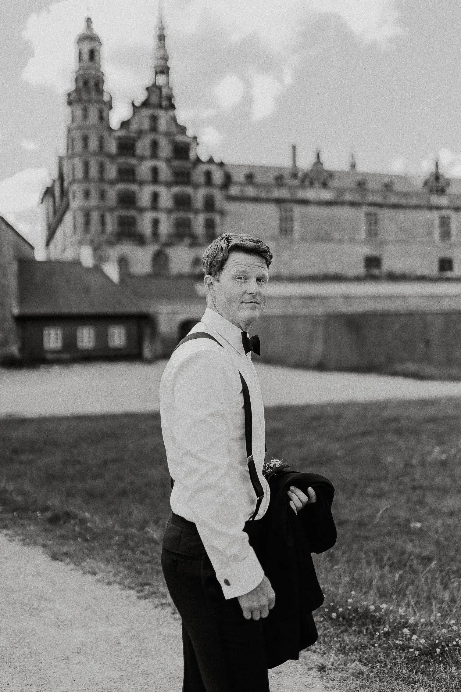 En mann i smoking holder en jakke og står på en plen foran historiske bygninger i Danmark, med overskyet himmel.