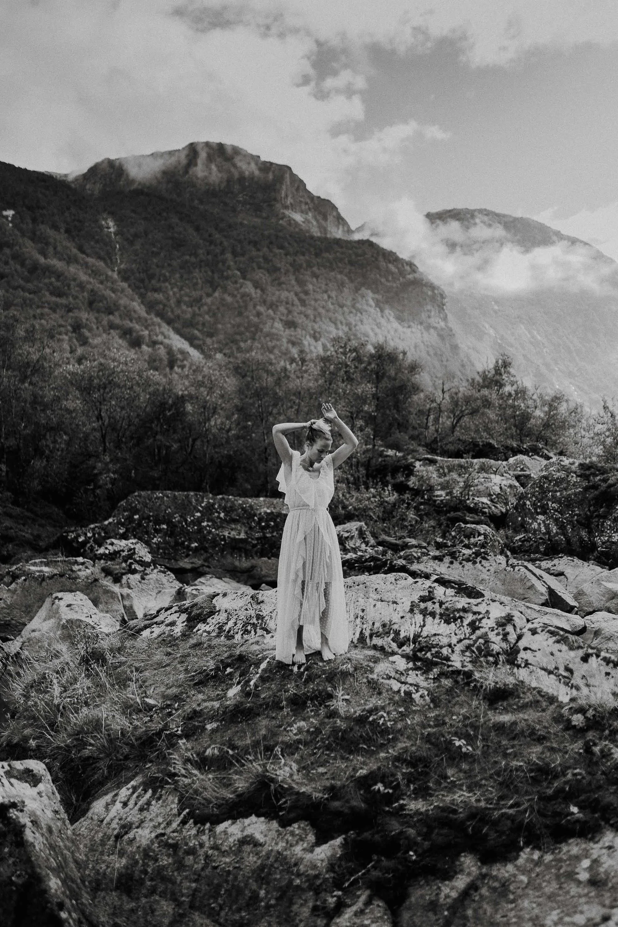 Sort hvitt bilde av en kvinne i flagrende kjole som står i steinete landskap omgitt av fjell og trær.