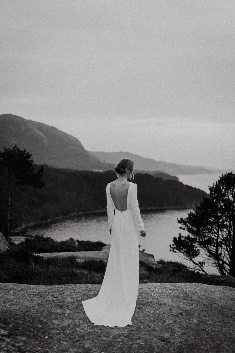 Et sort hvitt fotografi av en kvinne i lang hvit kjole som står på en steinete kant med utsikt over elv og fjell i det fjerne.