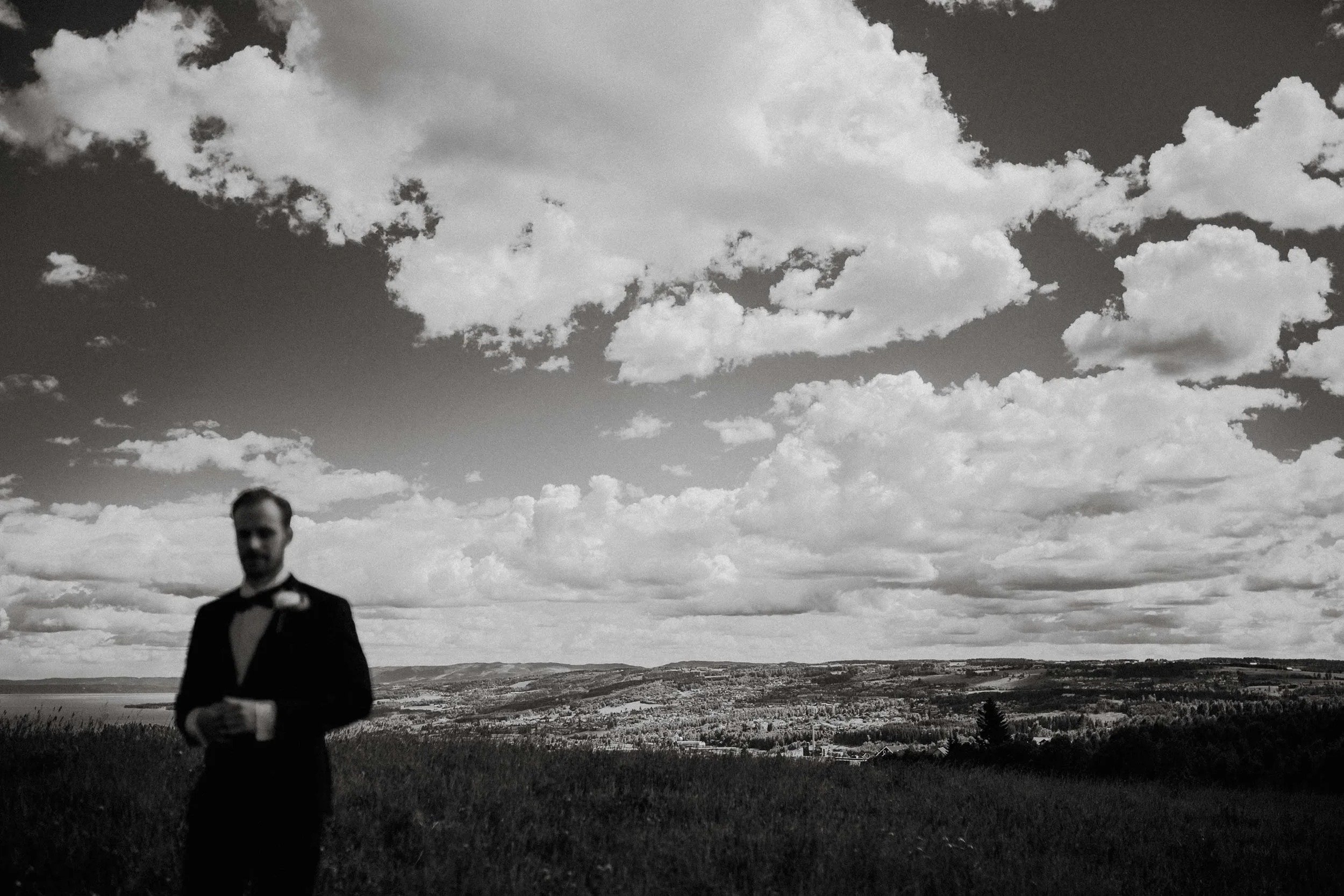 Uskarpt bilde av en mann i smoking som står utendørs foran et landskap med åser og skyer på himmelen.