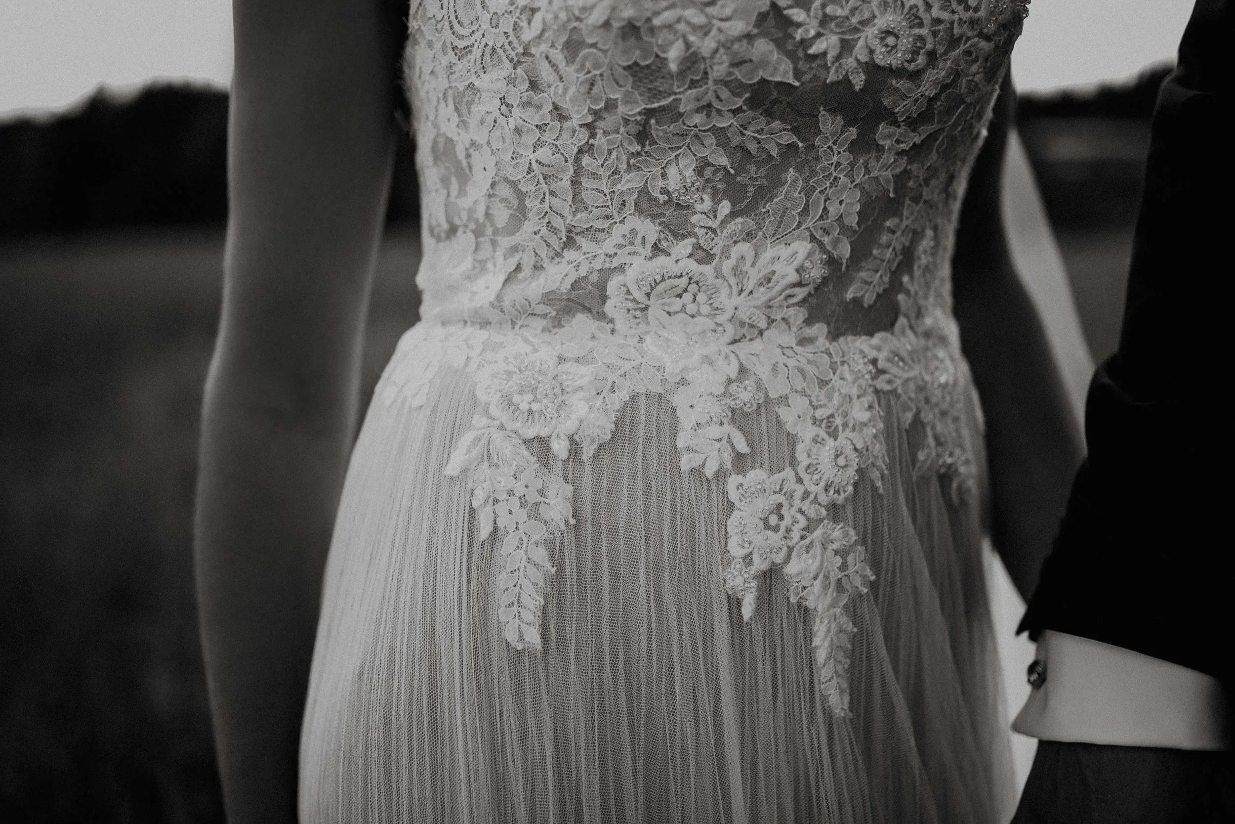 Nærbilde av en brudekjole med detaljert blondeoverdel og mykt fallende stoff. En arm og erme fra en annen person er synlig.