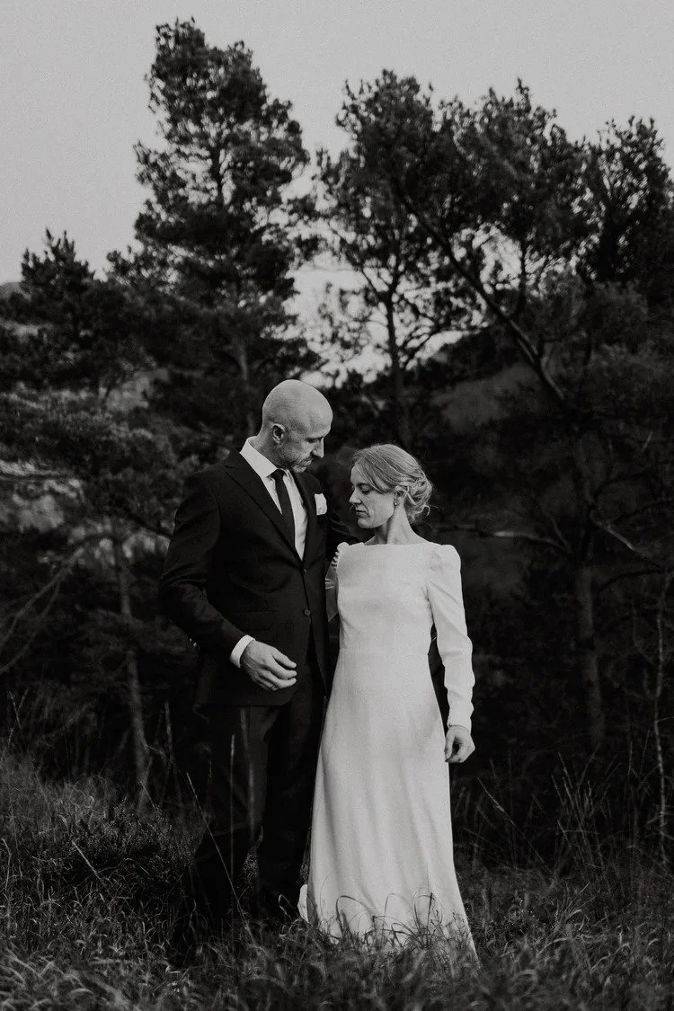 Sort hvitt bilde av et par som står utendørs i et naturlig miljø med trær i bakgrunnen. Mannen er iført dress og slips, kvinnen har lang hvit kjole. De ser ømt på hverandre.