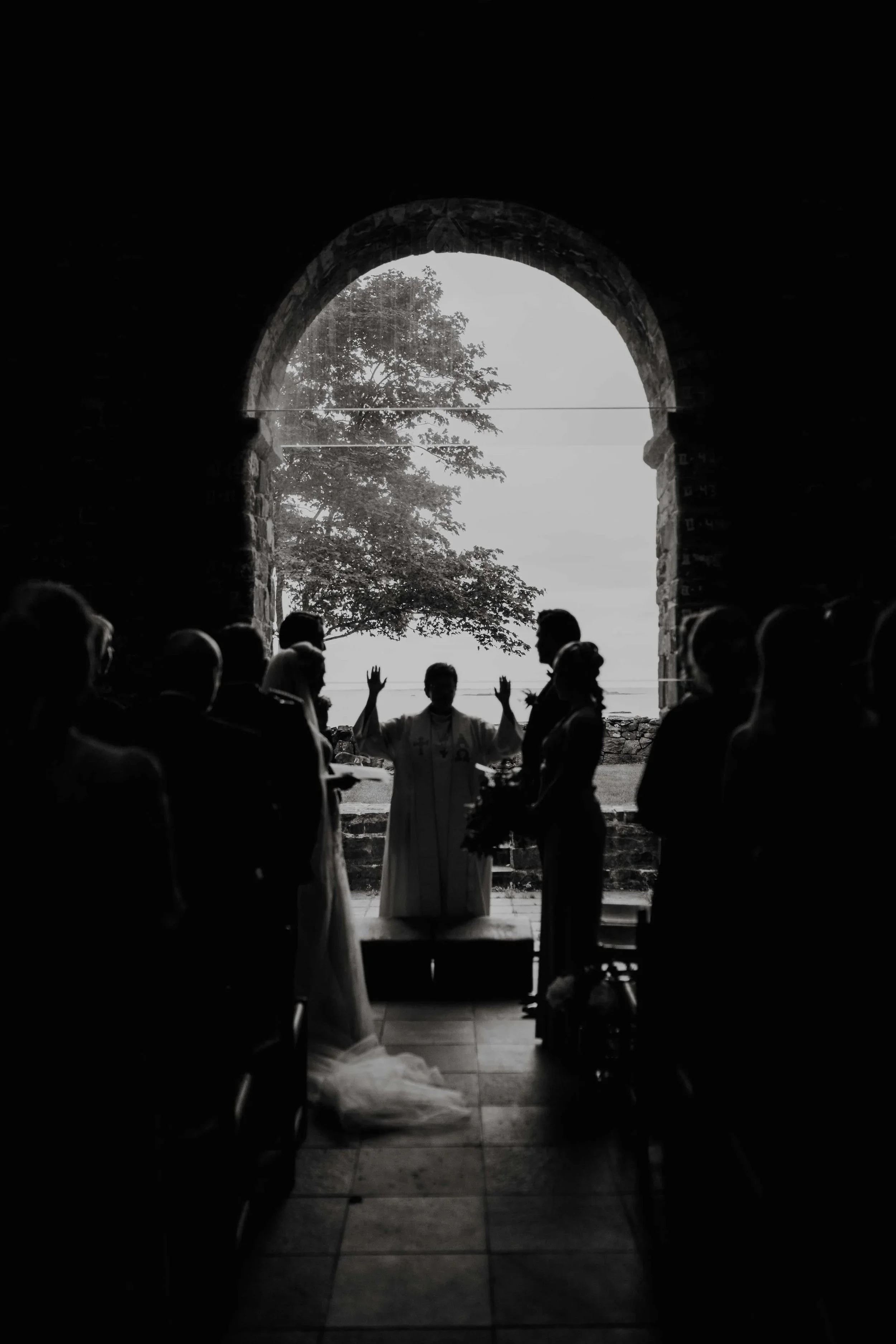Silhuett av en bryllupsseremoni inne i en steinbue, med vigsler som står utenfor buen med armene hevet, gjester sitter på hver side av midtgangen, med utsikt mot tre og åpen himmel.