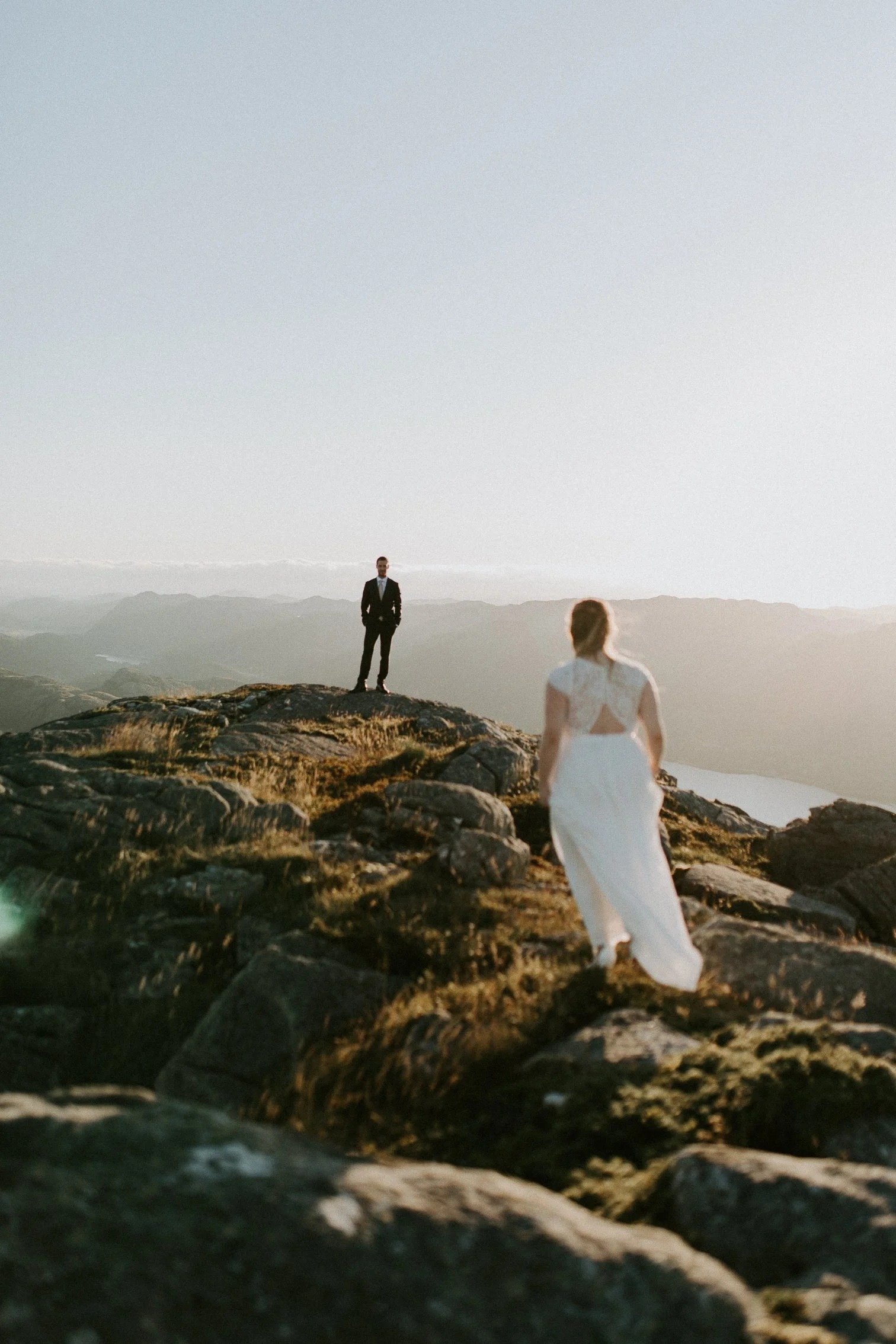 En kvinne i hvit kjole står på steiner og ser mot en mann i sort dress som står på en stein lenger unna, i et fjellandskap i solnedgang.