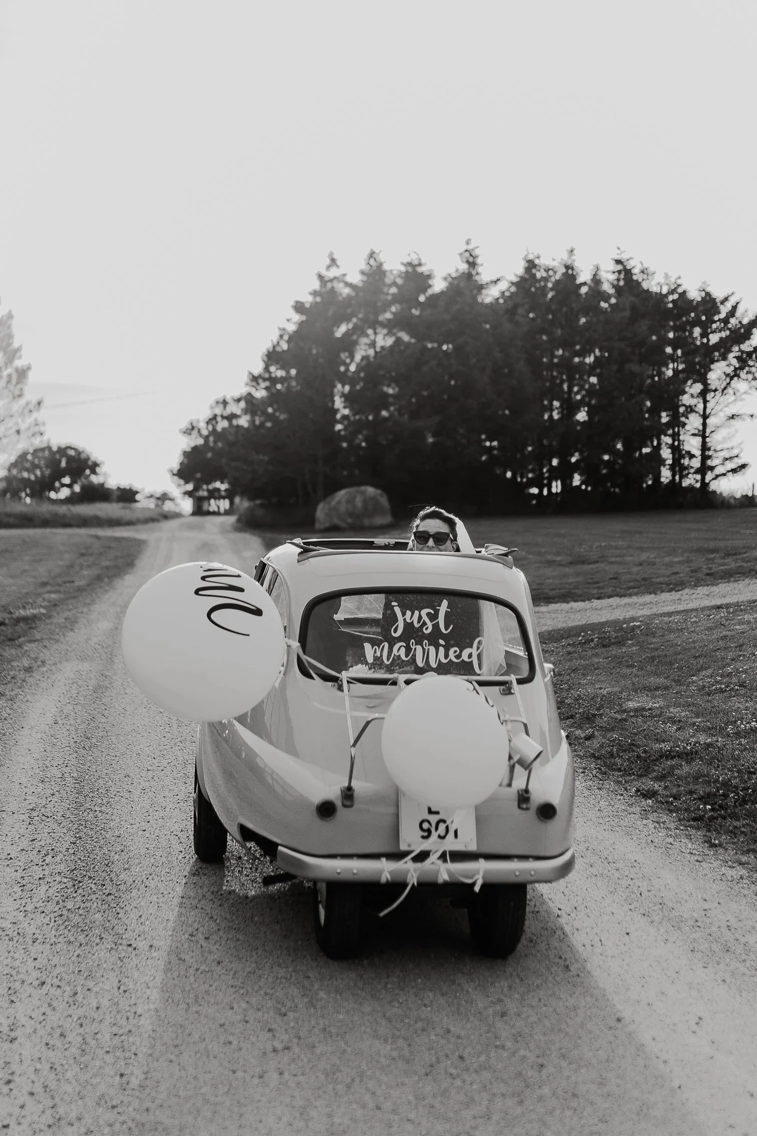En liten vintage mikrobil dekorert med ballonger og skilt med teksten just married, på en grusvei med trær i bakgrunnen.