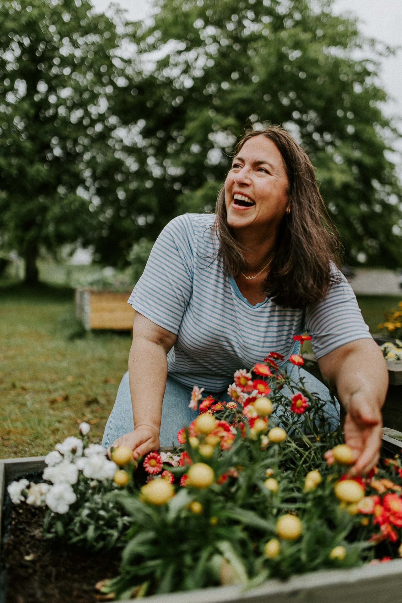 Kvinne som fikser i blomsterbed smiler og ser opp til venstre mens hun holder en hånd på blomster naturlig lys, reklamefotografering for stavanger kommune, områdesatsingen i hillevåg