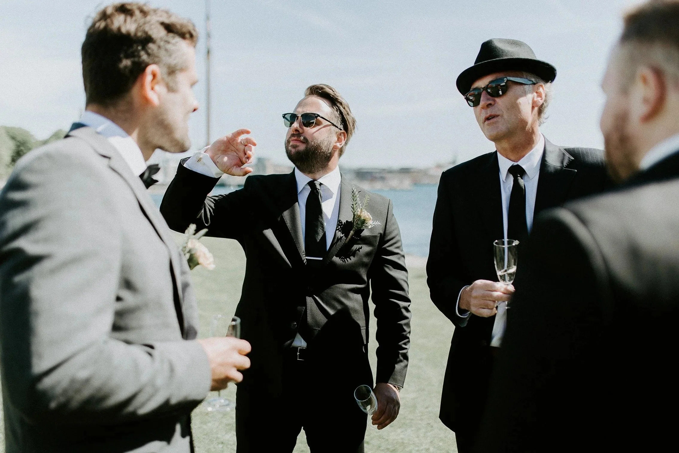 Menn i dresser og solbriller i samtale utendørs nær vann.