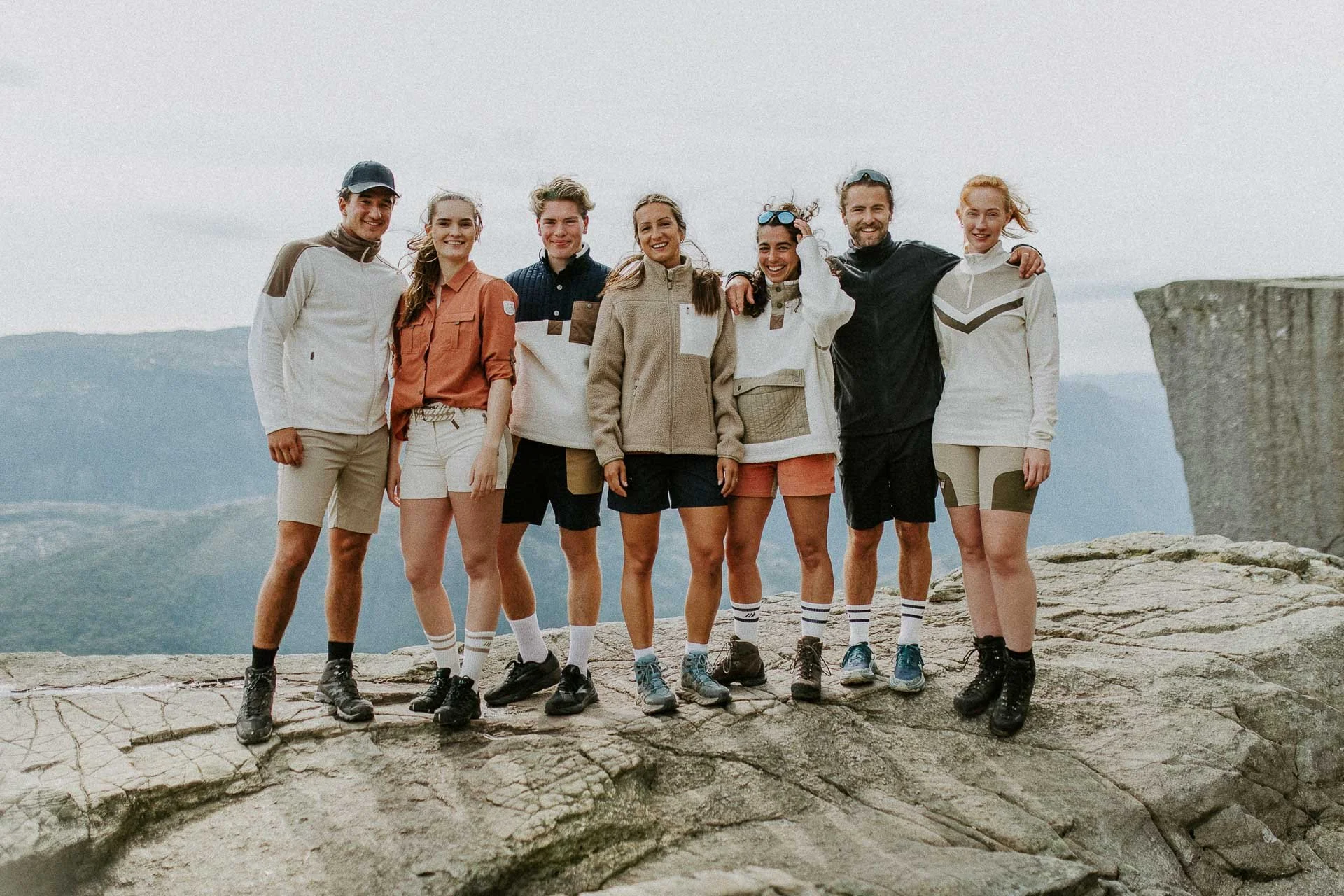 Gruppebilde tatt på toppen med Preikestolen i bakgrunnen alle ser mot kamera hun ene holder hånd opp mot solbriller lett skygge fra beina deres alle smiler Reklamefotografering av modeller til kampanje for Skogstad bilder tatt i preikestolenområdet b