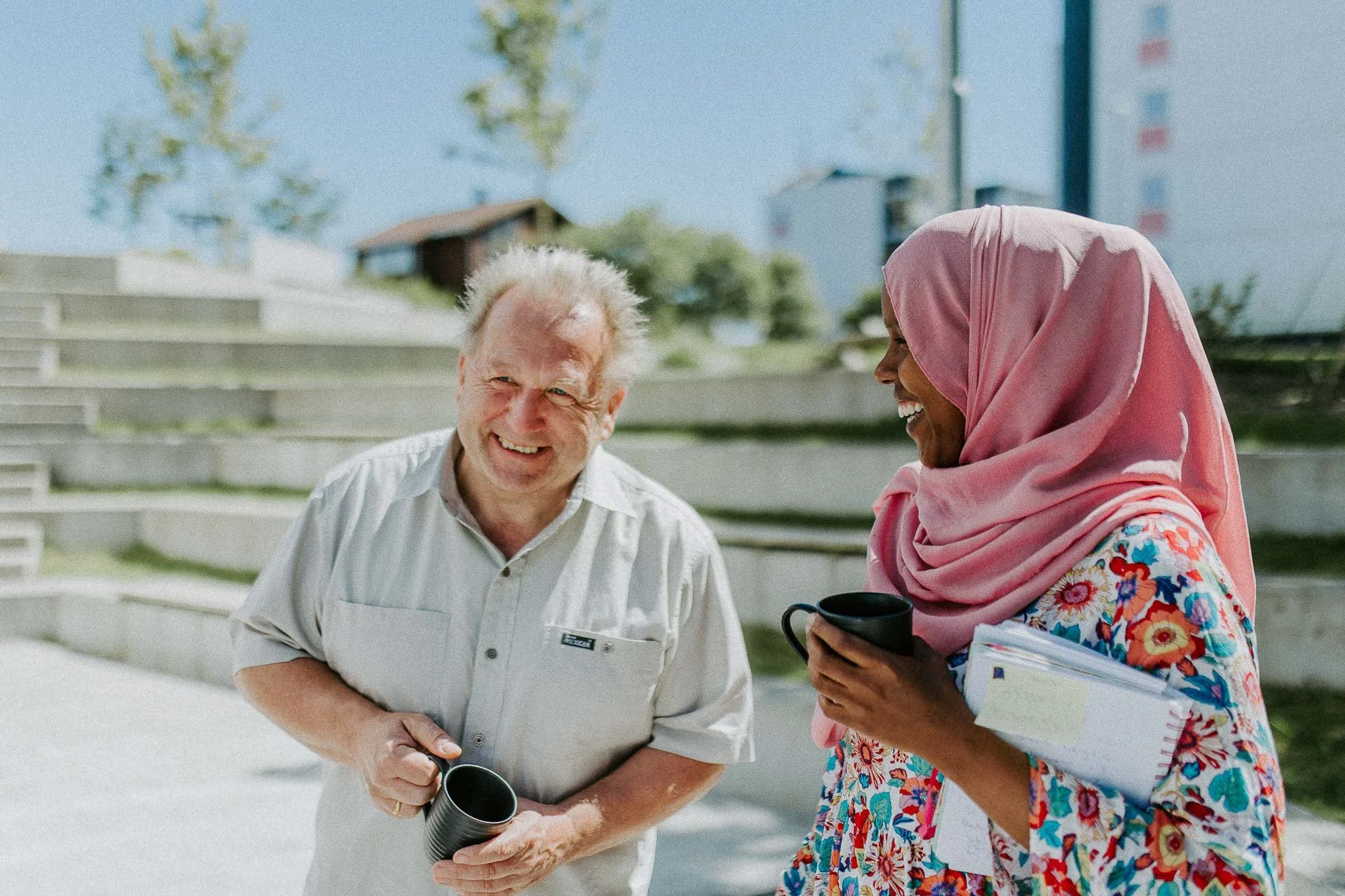 To personer i samtale Mann og kvinne begge ler og holder kaffekopp trapp i bakgrunnen naturlig lys, reklamefotografering for stavanger kommune, områdesatsingen på kvernevik tatt på sommeren