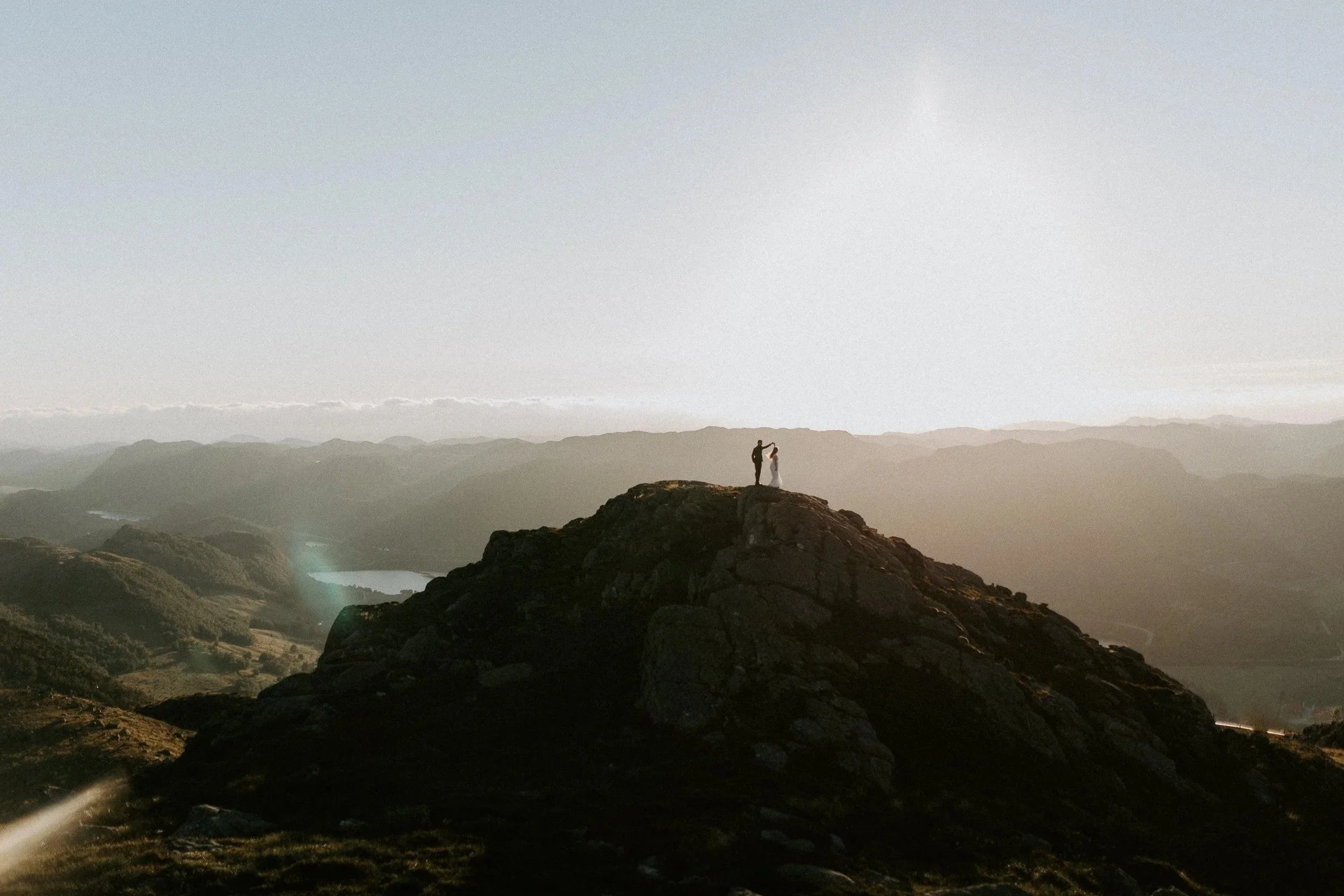Et par står på toppen av et steinete fjell, holder hender og vender seg mot hverandre i solnedgang, med utsikt mot bølgende åser og fjerne fjell.