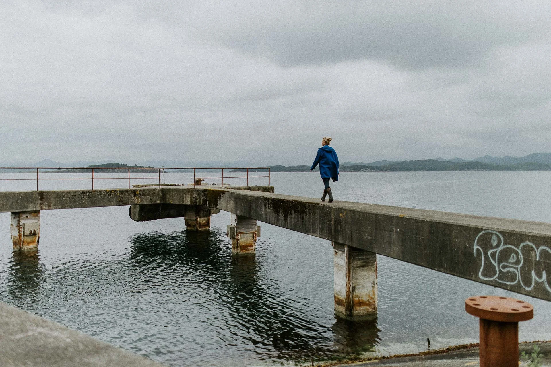 Kvinne med blå jakke går over på grå betong over havet naturlig lys, reklamefotografering for stavanger kommune, områdesatsingen i hillevåg fjell og sjø og horisont