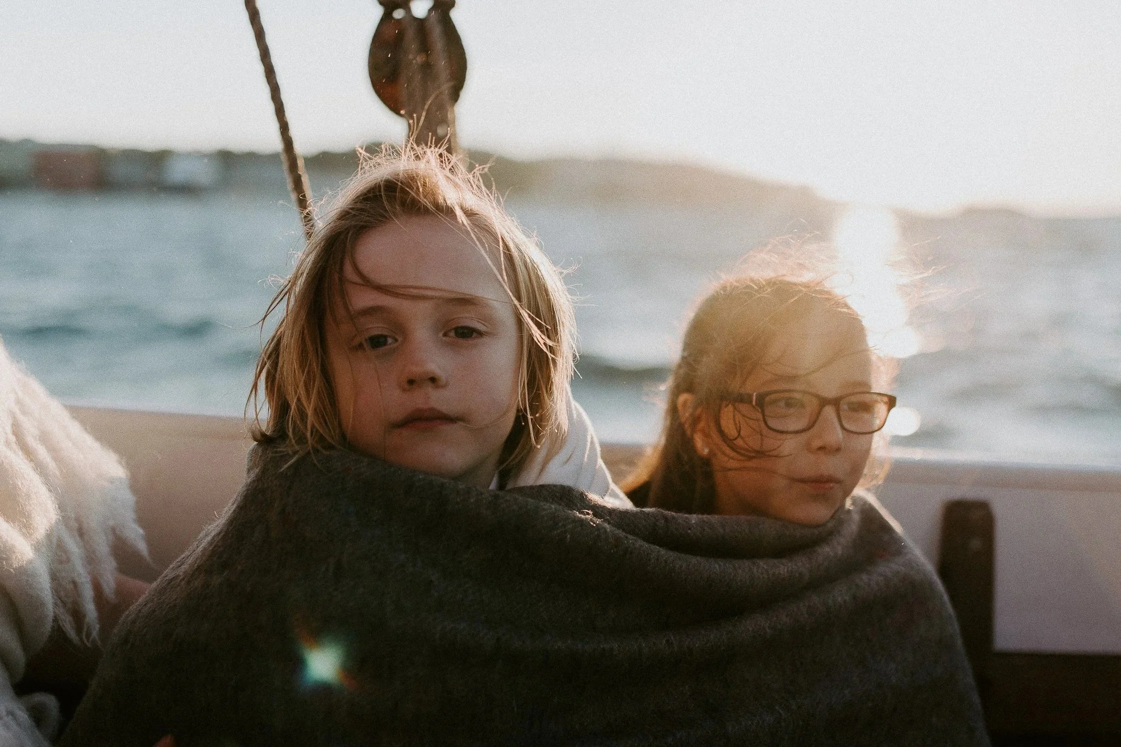 To barn, en jente med briller og en gutt, sitter pakket inn i et teppe på en båt i solnedgang, med vann og land i det fjerne.