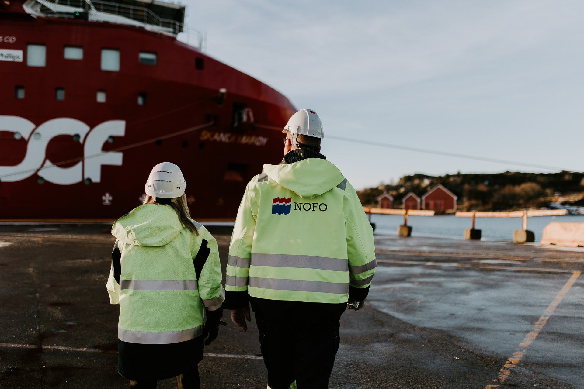 Bilde tatt utendørs to personer med rygg mot kamera gul arbeidsjakke med Nofo logo på sjø og båt i bakgrunnen reklamefotografering for nofo norsk oljevernberedskap tatt i naturlig lys