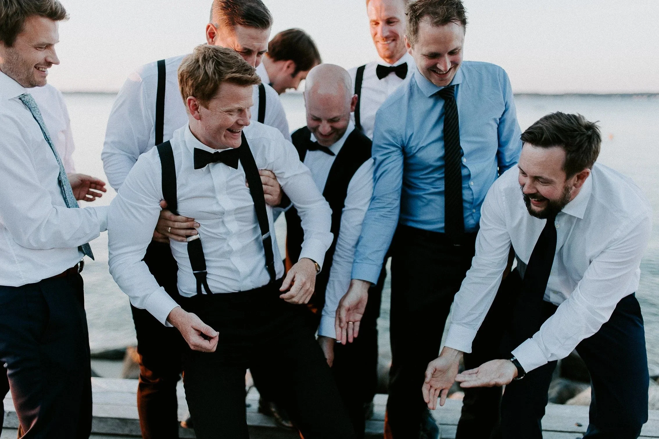 En gruppe menn i formelt antrekk med hvite skjorter, bukseseler og slips ler og har et øyeblikk sammen på en brygge ved vannet.