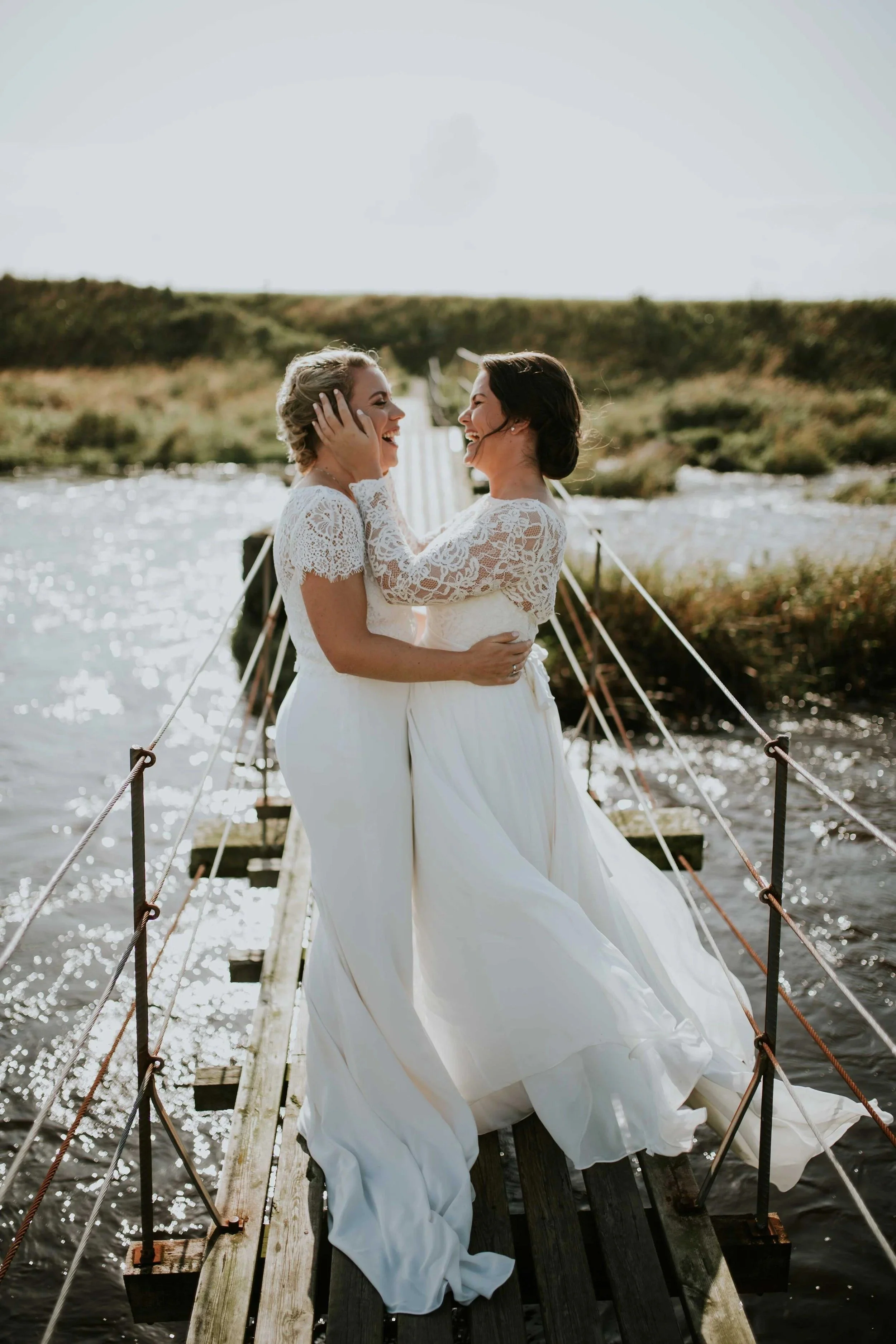 To kvinner i brudekjoler klemmer hverandre og smiler på en trebro over vann utendørs.
