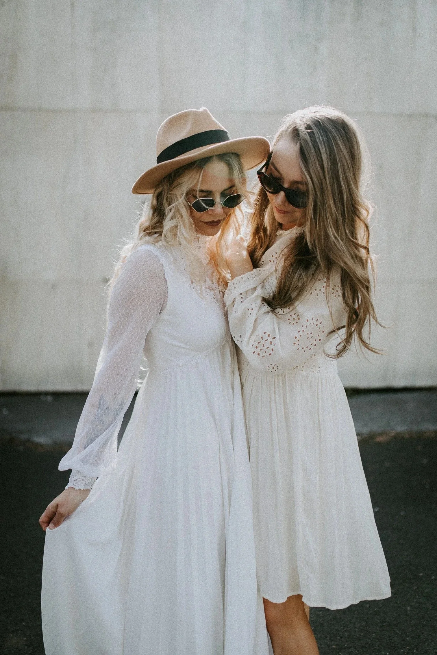 Brudepar to kvinner i hvite kjoler og solbriller står tett sammen utendørs. Den ene har en bredbremmet hatt med sort bånd.
