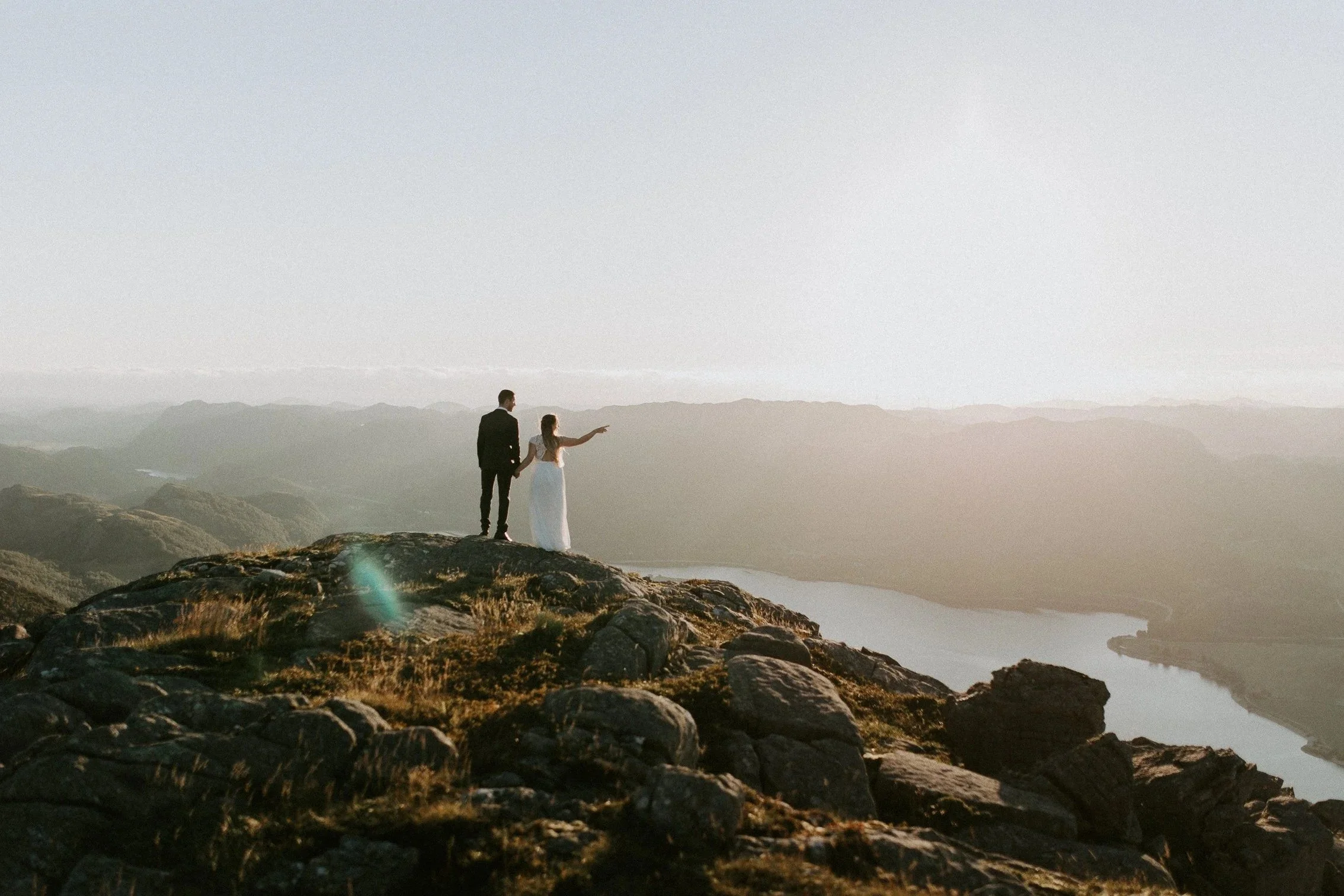 Et brudepar står på en steinete høyde med utsikt over elv og fjellandskap i solnedgang, holder hender mens bruden peker mot landskapet.