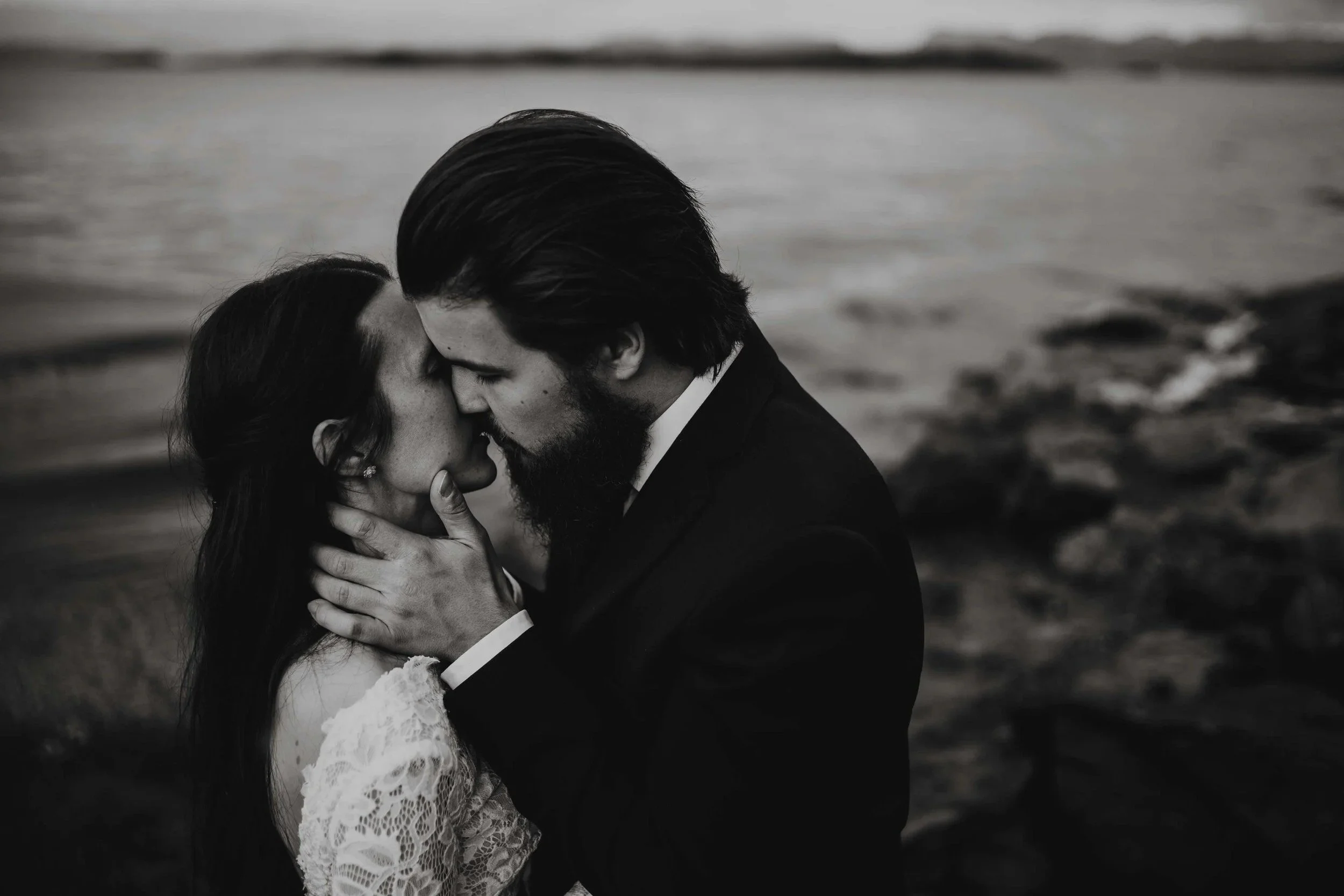 Sort hvitt bilde av et brudepar som kysser hverandre utendørs nær vann.