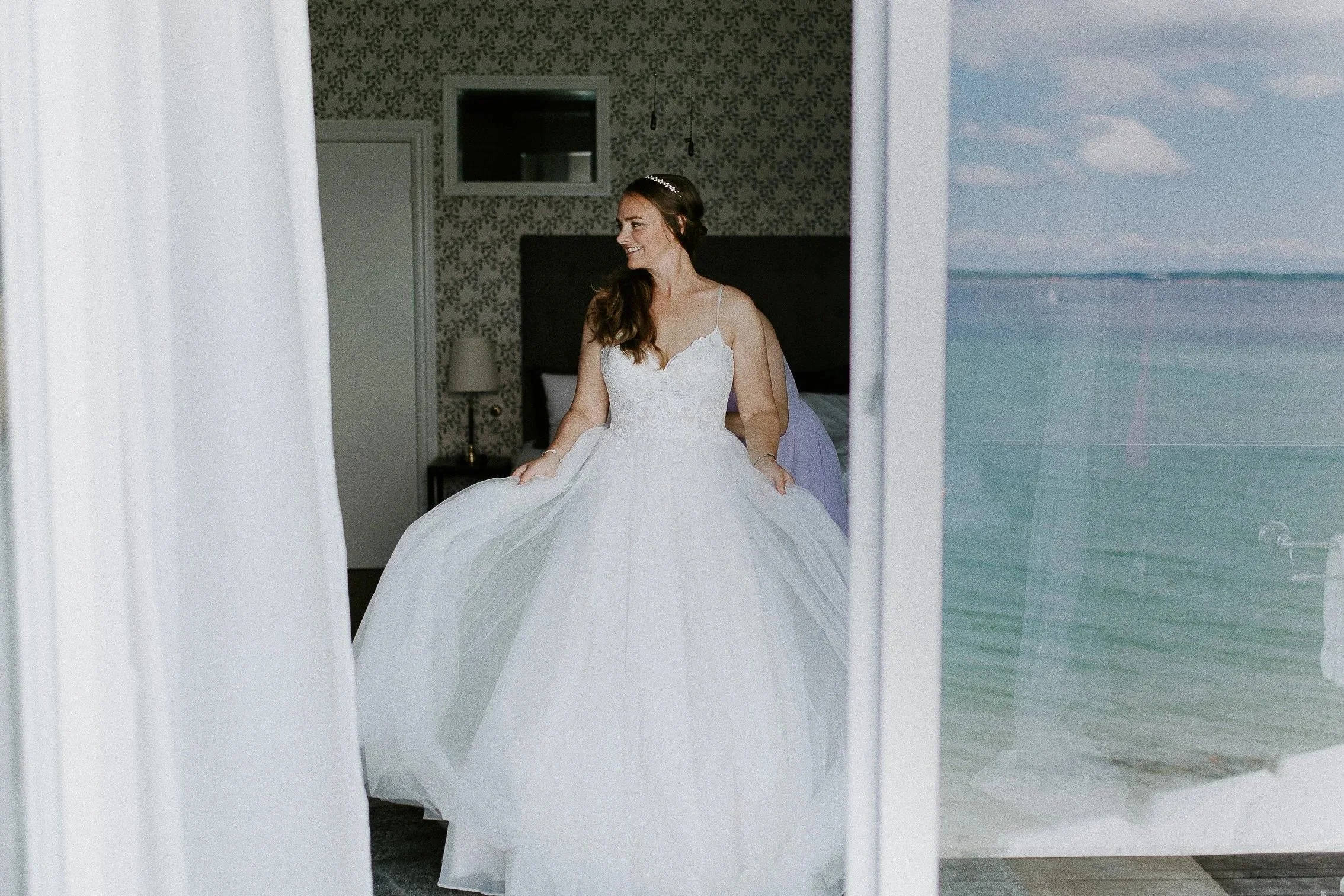 En brud i brudekjole sitter på en seng, sett gjennom et vindu eller glassdør. I refleksjonen ser vi blå himmel horisont hav