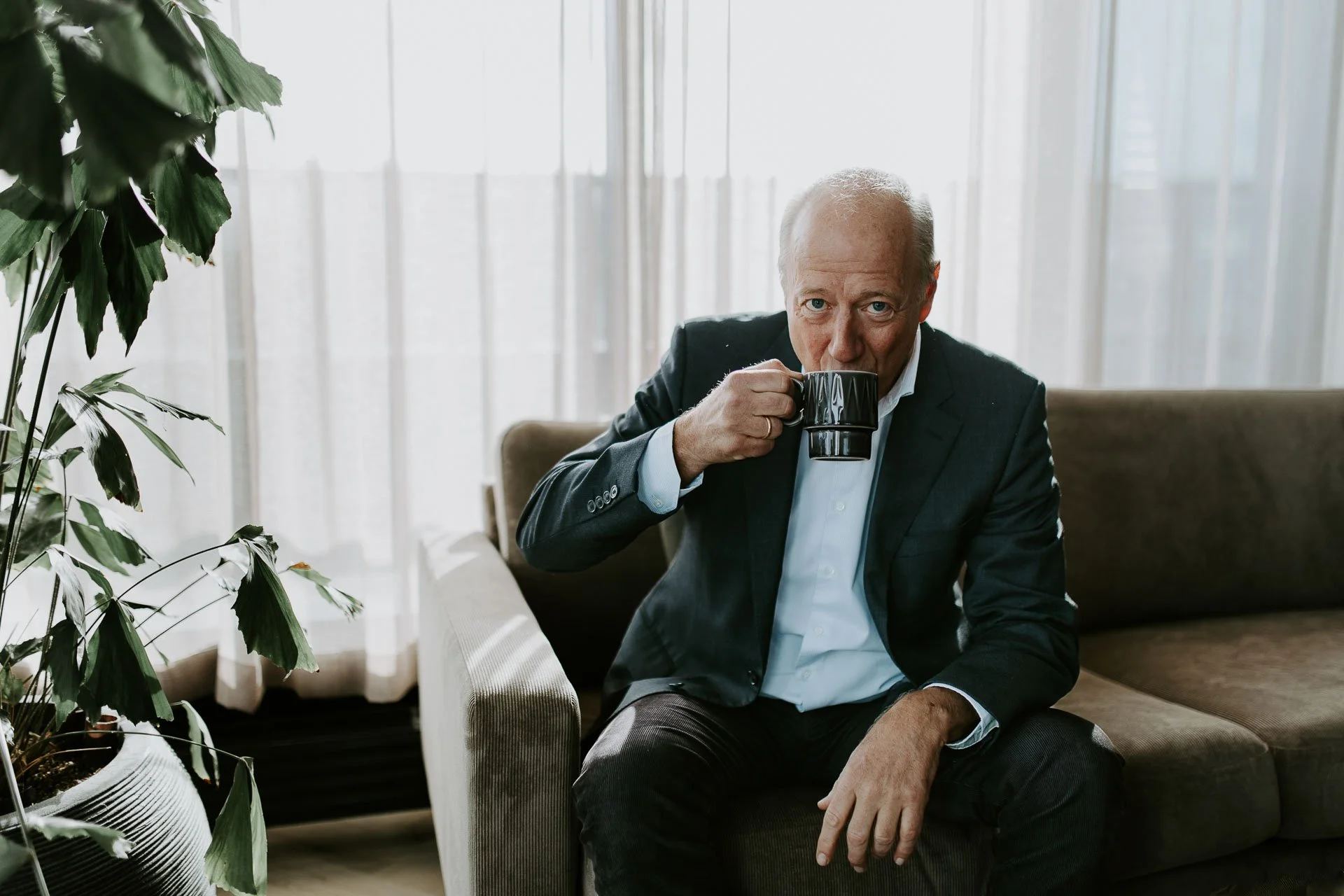 Portrett av direktør i norsk Oljevernberedskap nofo drikker en kopp med kaffe ser i kamera har på dress og skjorte sitter i sofaen reklamefotografering for nofo norsk oljevernberedskap tatt i naturlig lys