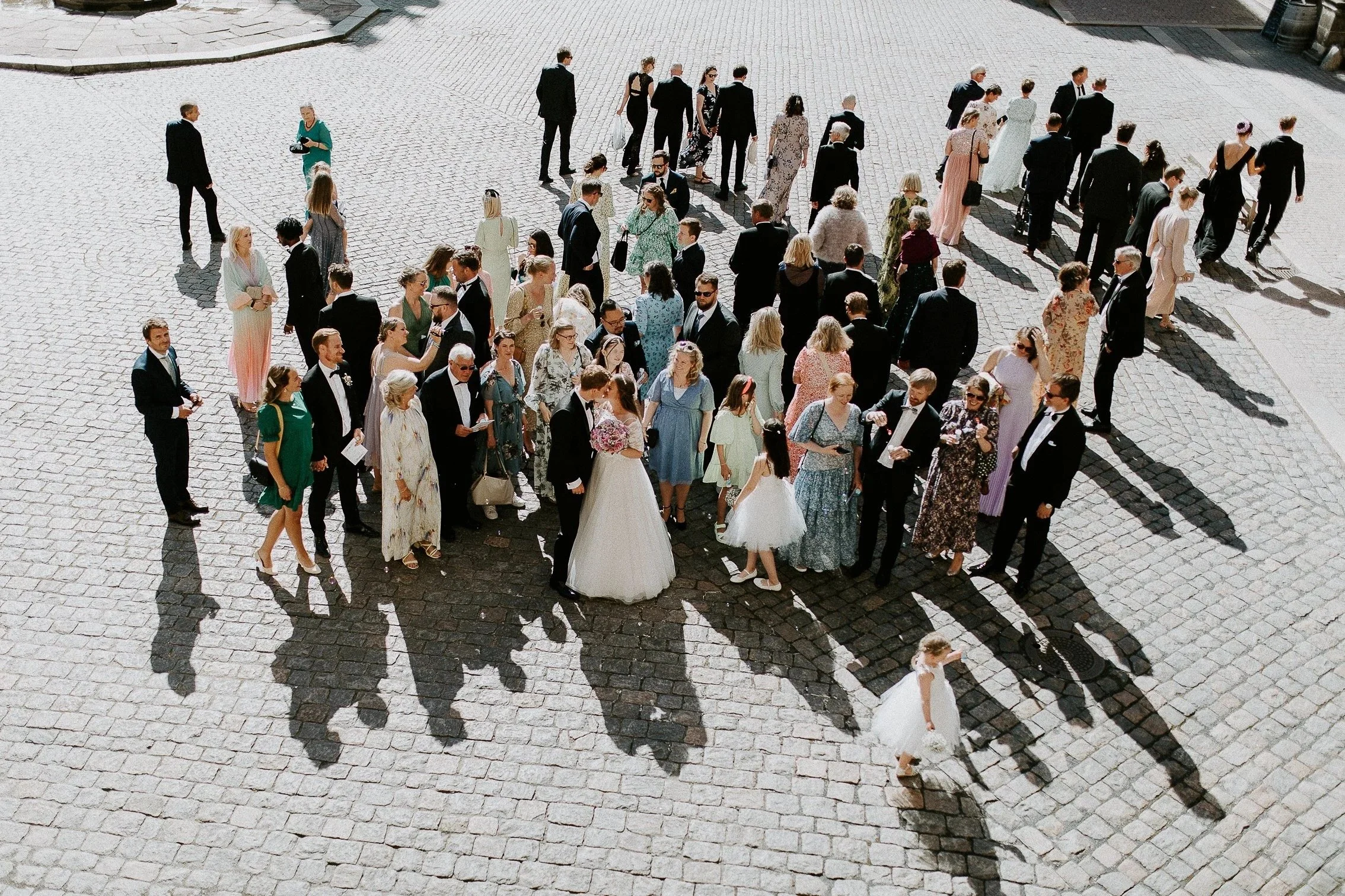 En gruppe bryllupsgjester står i en brosteinsgate, med brudeparet i sentrum, og lange skygger i sollys.