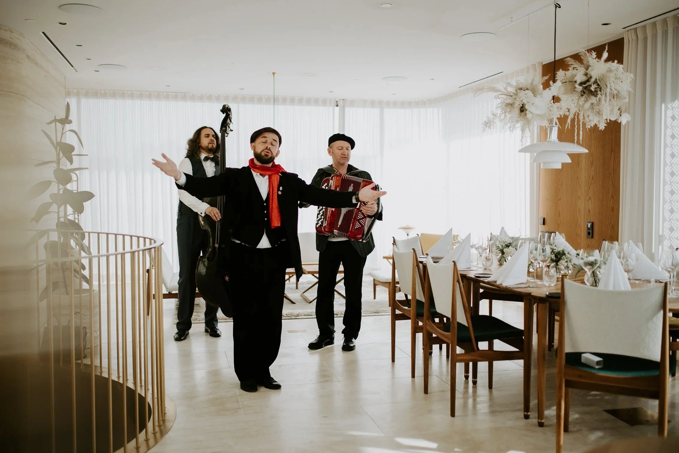 Et band med tre musikere opptrer i en elegant restaurant med hvite gardiner og treinteriør. Vokalisten står fremst med armene ut, akkompagnert av bass og trekkspill.