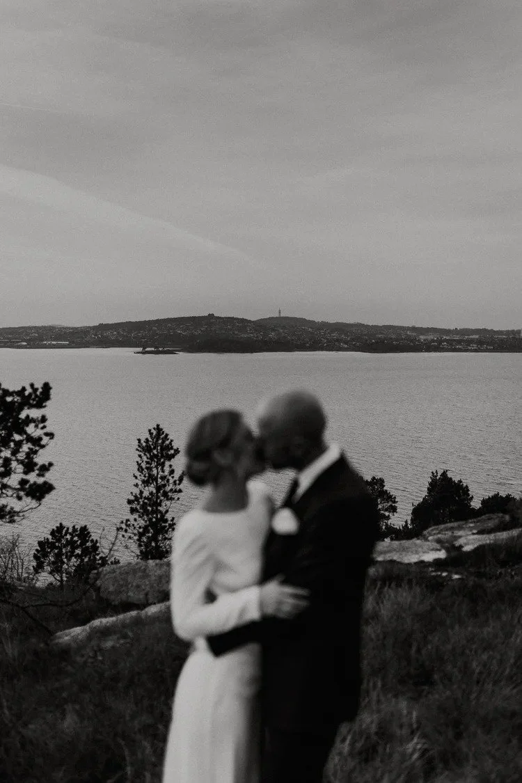 Et uskarpt sort hvitt bilde av et brudepar som omfavner hverandre utendørs nær vann, med trær og landskap i bakgrunnen.