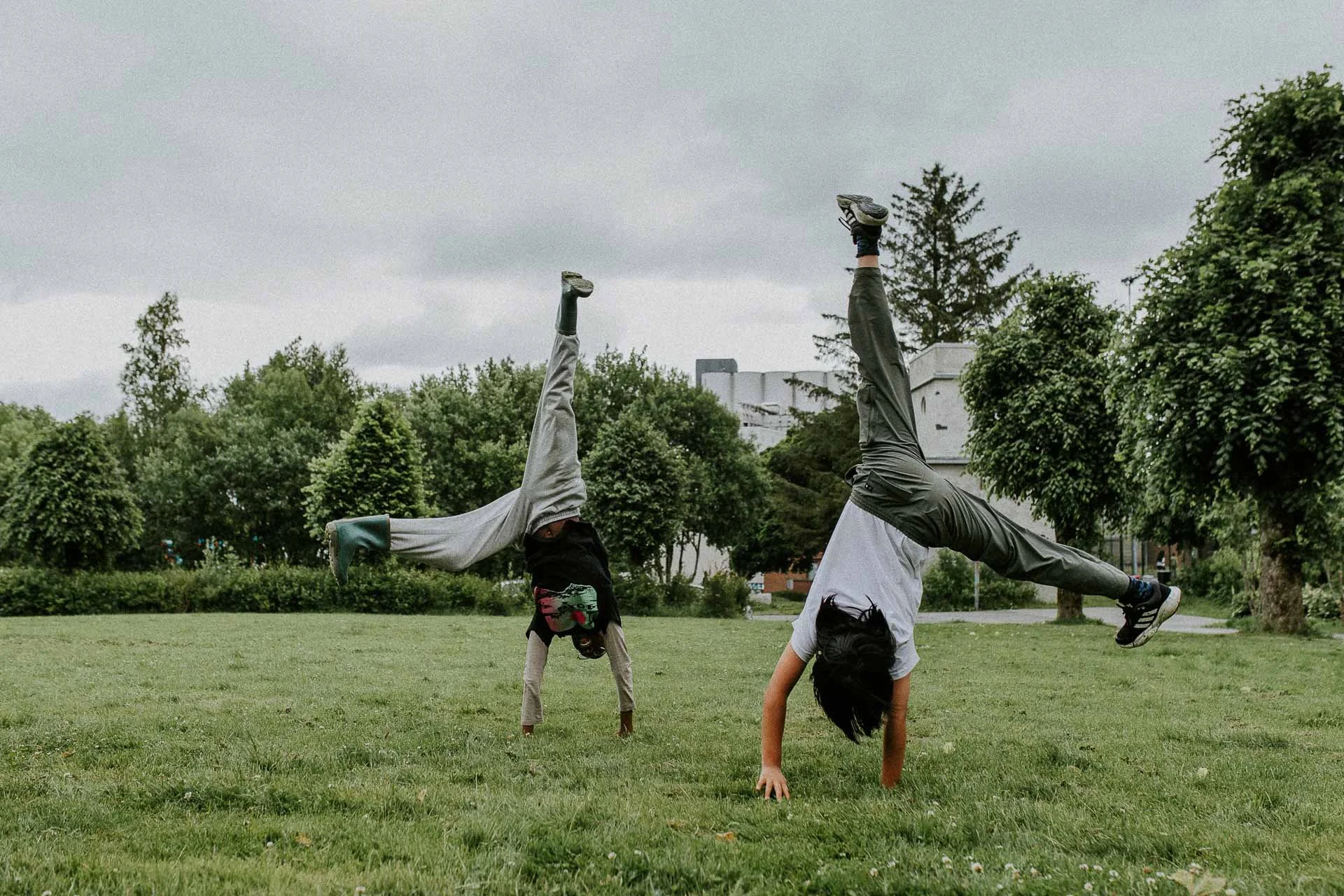 To barn som slår hjul på gressflate man kan se trær og bygge Bakgrunn det er blant annet siloen på Hillevåg naturlig lys, reklamefotografering for stavanger kommune, områdesatsingen i hillevåg