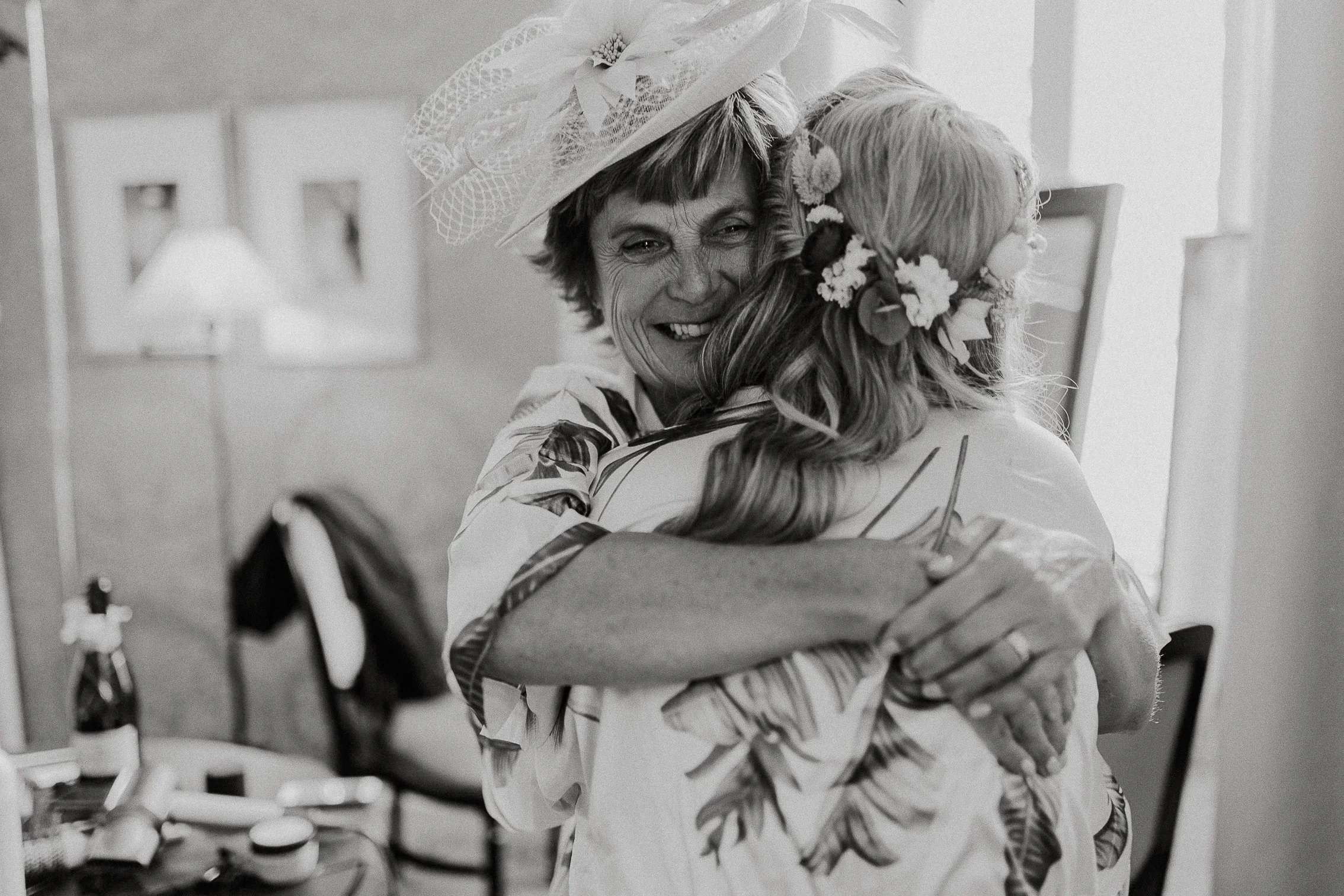 En kvinne i blomstrete kjole og en annen kvinne med blomsterkrans klemmer hverandre og smiler.