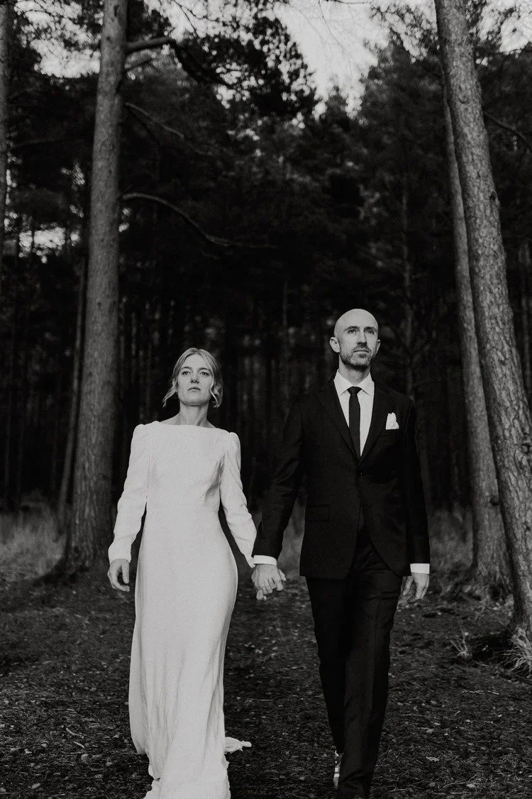 Sort hvitt bilde av et brudepar som går hånd i hånd gjennom skog. Kvinnen har lang hvit kjole, mannen er i sort dress og slips.
