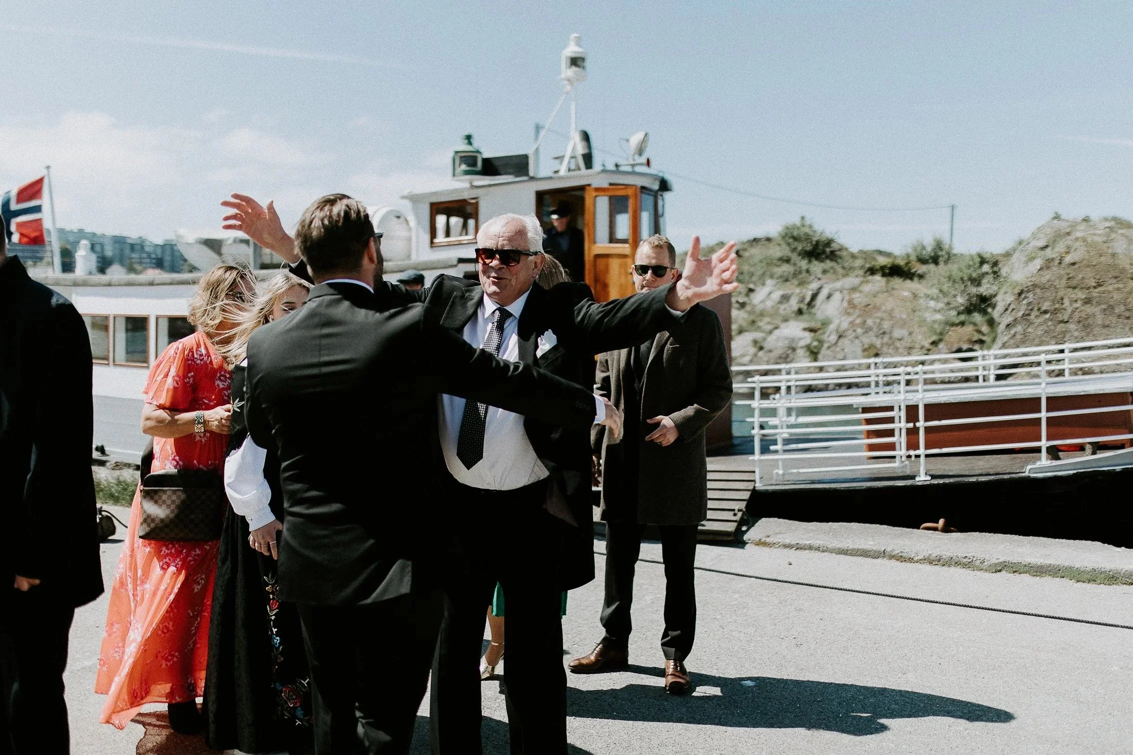 En gruppe mennesker i formelt antrekk står ved en brygge med båt i bakgrunnen. En mann gestikulerer mens han snakker.
