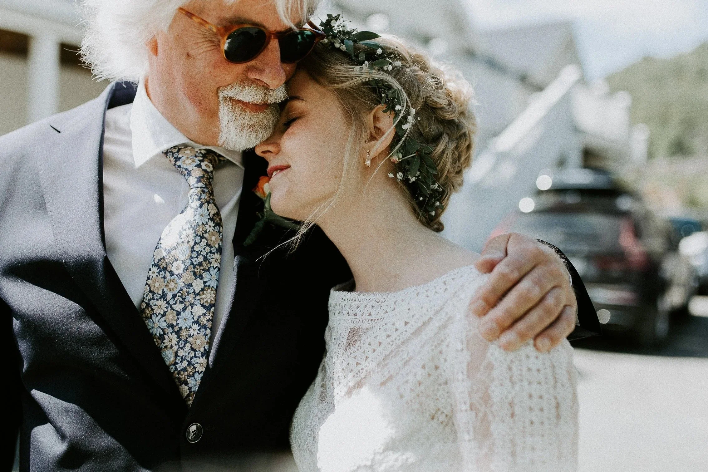 En eldre mann med solbriller klemmer en yngre kvinne i hvit blondekjole. De deler et nært og gledelig øyeblikk utendørs.
