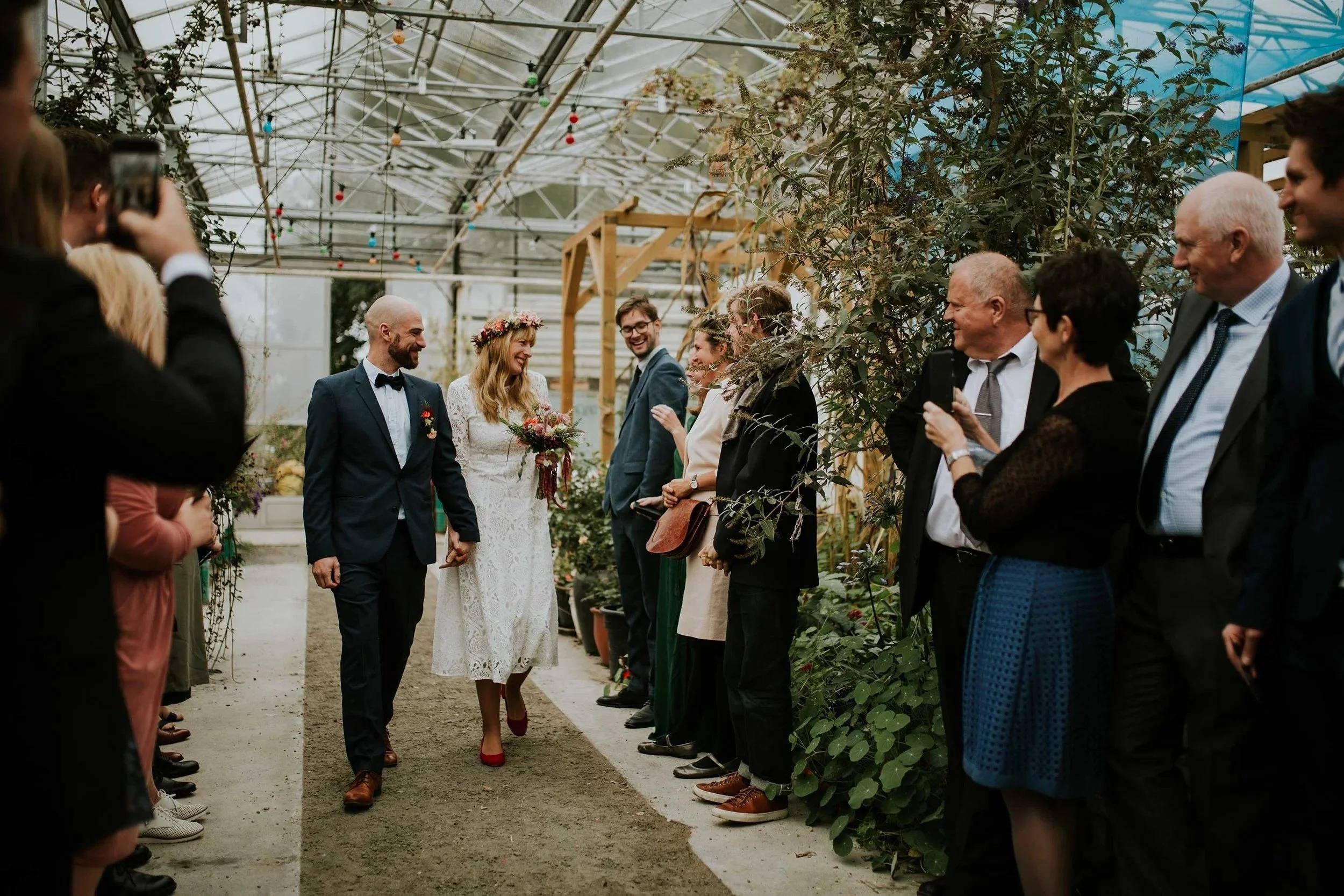Bryllupsseremoni i et drivhus der brud og brudgom går ned midtgangen. Gjester smiler og tar bilder, omgitt av planter og glasstak.