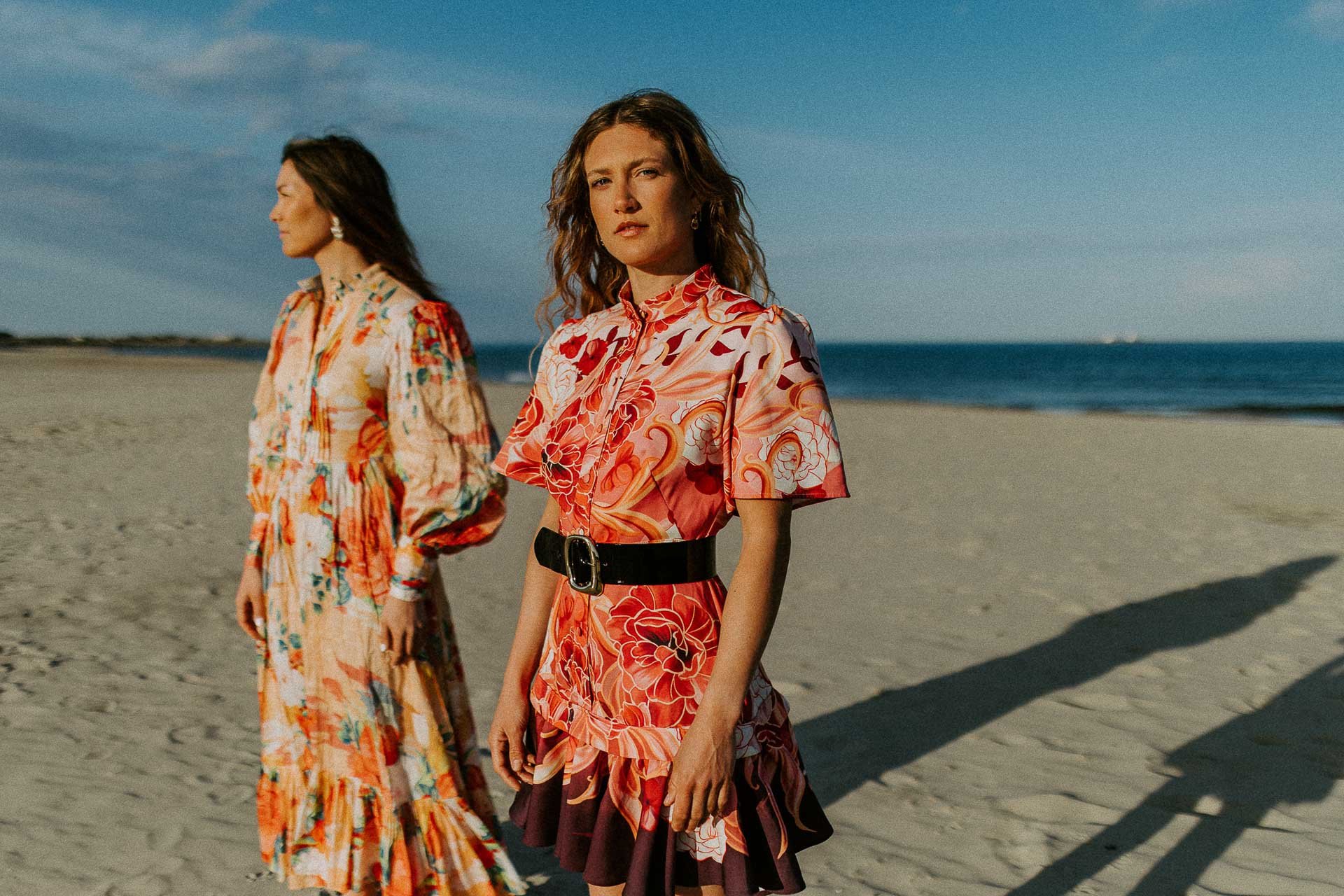 To kvinner i fargerike blomstrete kjoler står på en sandstrand med havet i bakgrunnen under blå himmel.