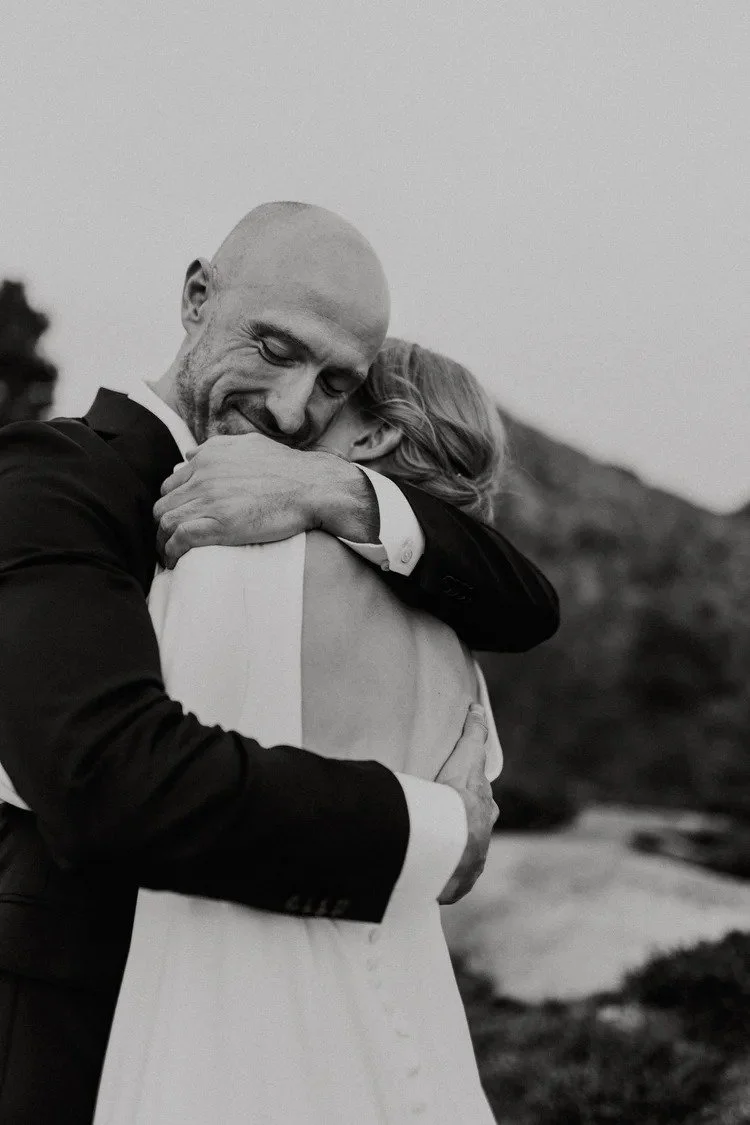 Sort hvitt bilde av en mann og en kvinne som klemmer hverandre tett og smiler, utendørs med fjell i bakgrunnen.