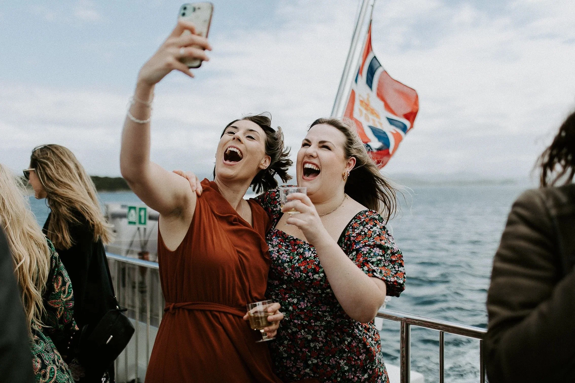 To kvinner på en båt tar selfie, smiler og ler. Den ene holder en drink, den andre et glass, med britisk flagg og havet i bakgrunnen.