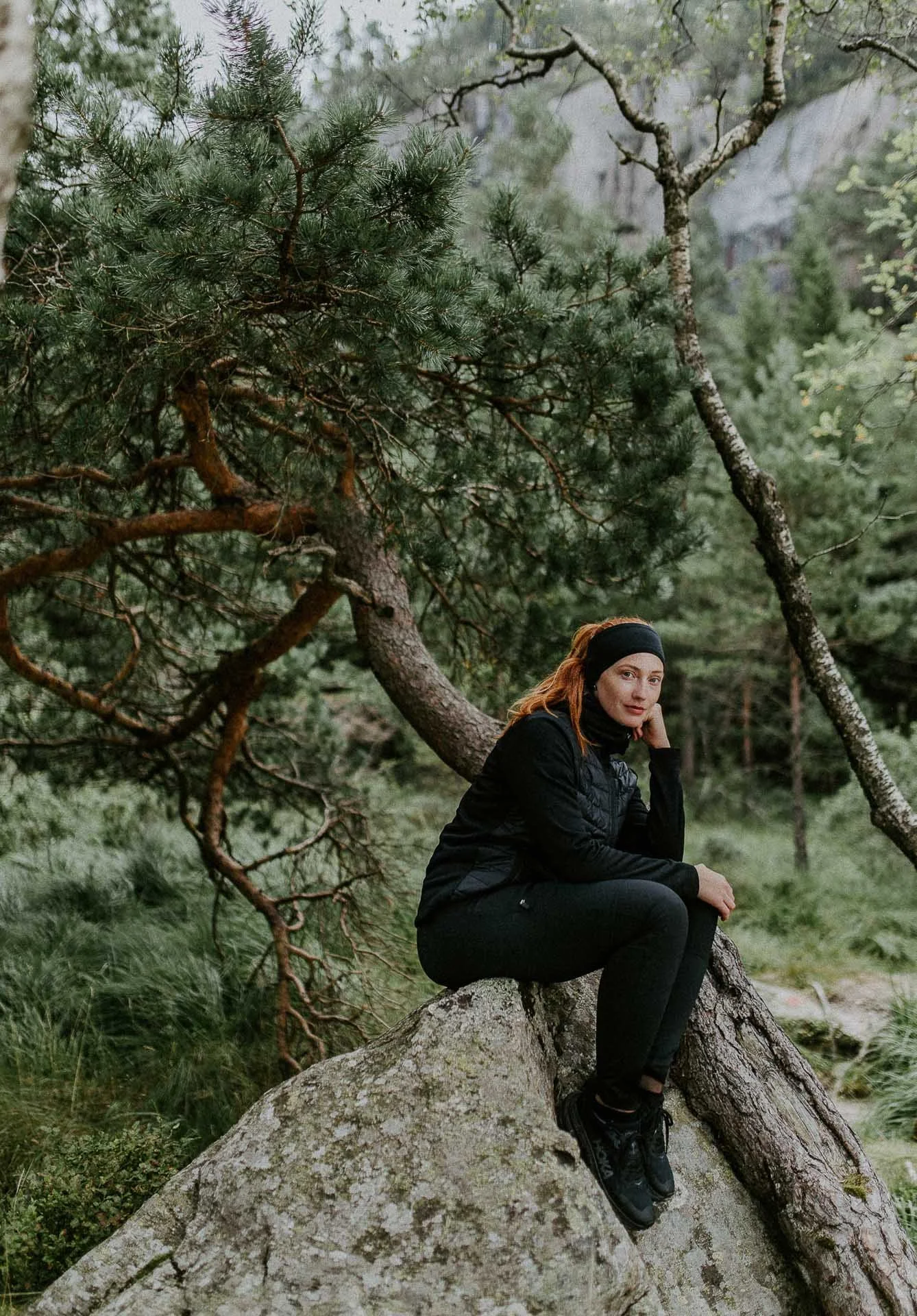 Kvinne sitter på stein ser mot kamera helfigur tre og skog i bakgrunnen hun har på sorte klær Reklamefotografering av modeller til kampanje for Skogstad bilder tatt i preikestolenområdet bilder av turklær