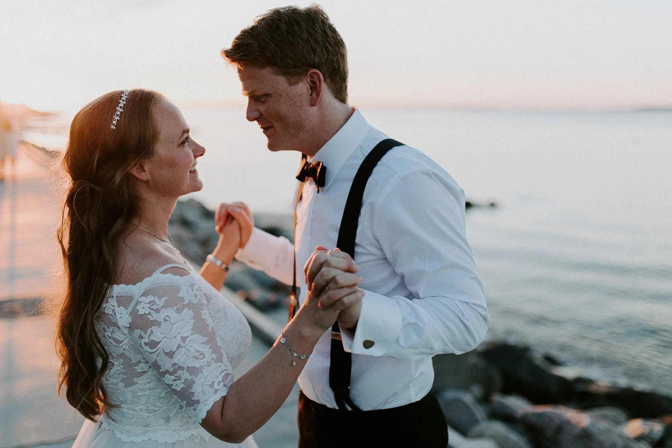 Et par danser ved vannkanten i solnedgang. Kvinnen i hvit blondekjole, mannen i hvit skjorte med bukseseler og sløyfe.