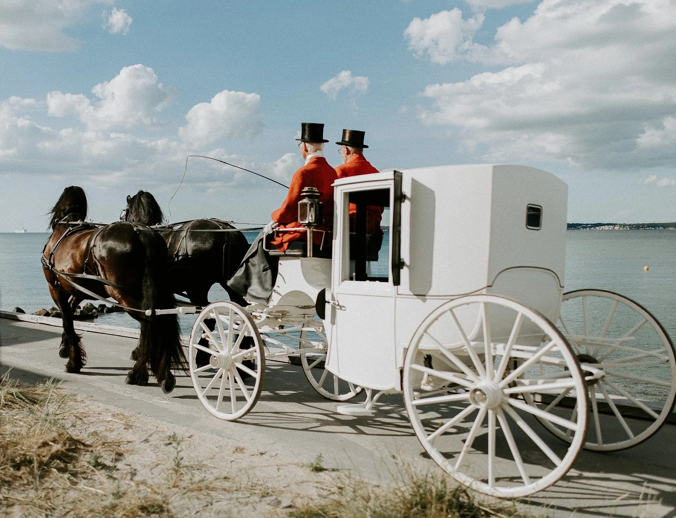 En hvit hestetrukket vogn med to kusker i røde jakker og sorte hatter langs vannkanten under blå himmel.