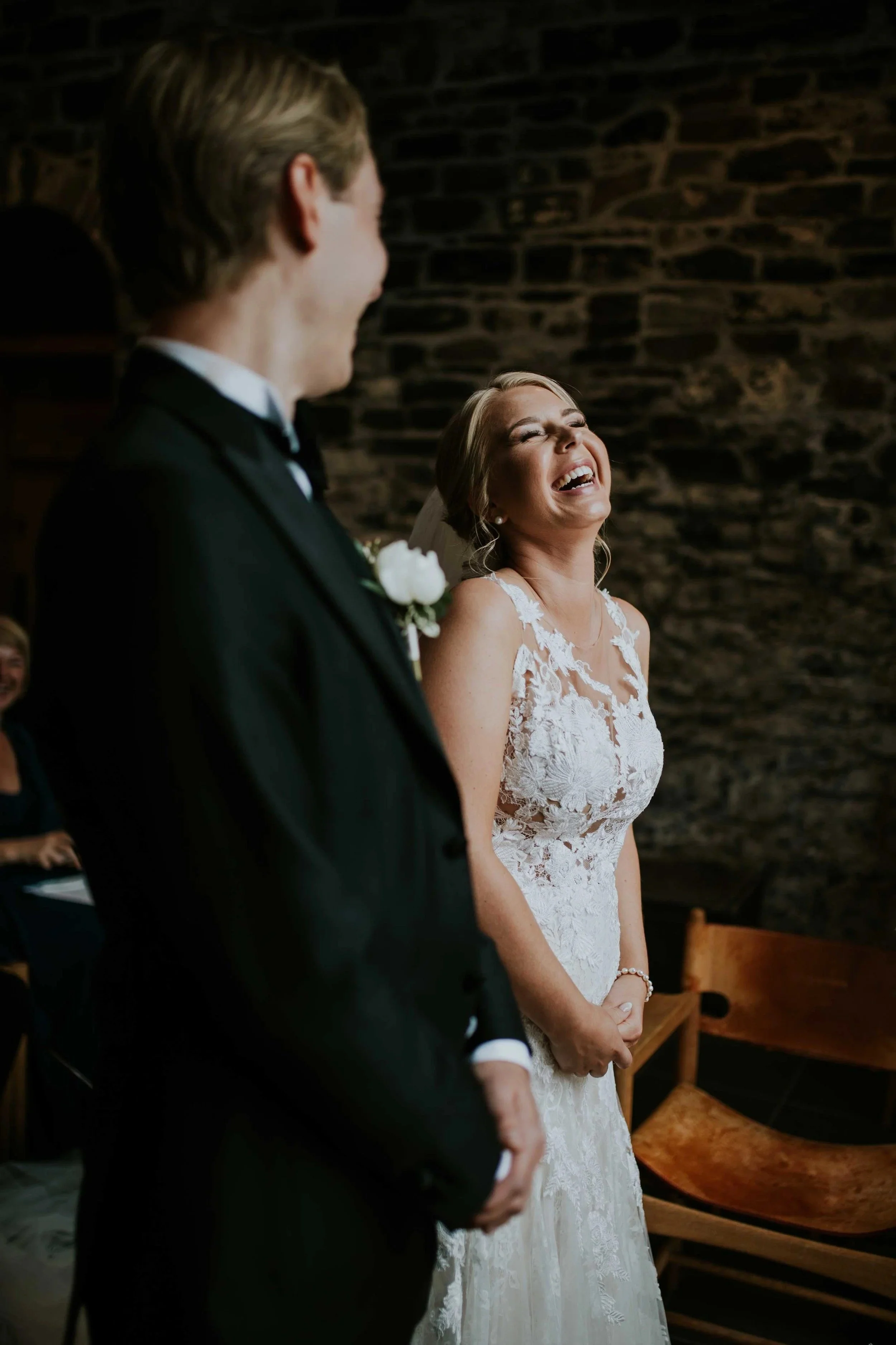 En brud ler under bryllupsseremonien, står ved siden av brudgommen i sort smoking med hvit brystblomst, foran en rustikk steinvegg.