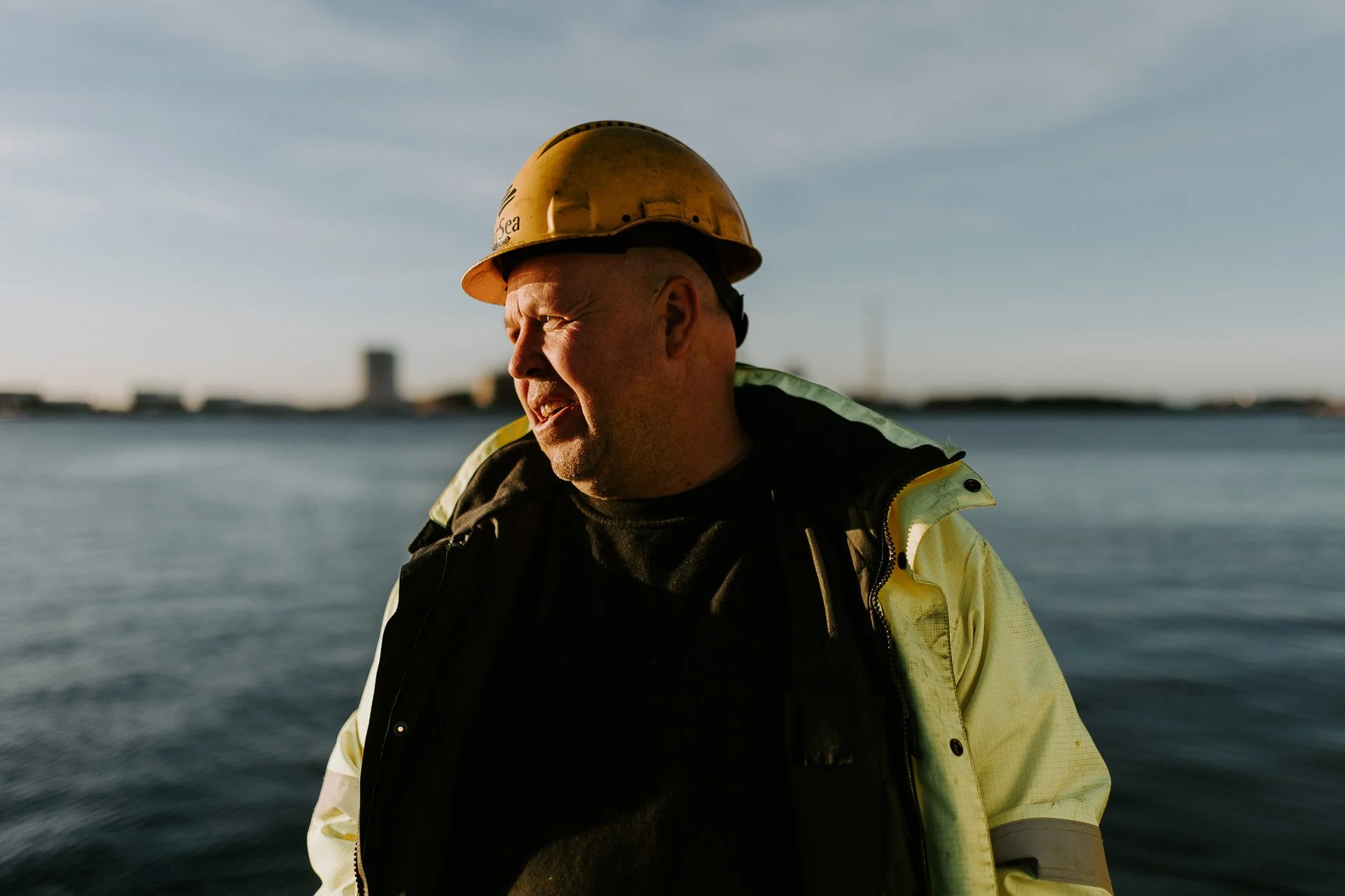 Fortsett av en mann som ser til siden har på hjelm og gul arbeidsjakke sjø i bakgrunnen reklamefotografering for nofo norsk oljevernberedskap tatt i naturlig lys