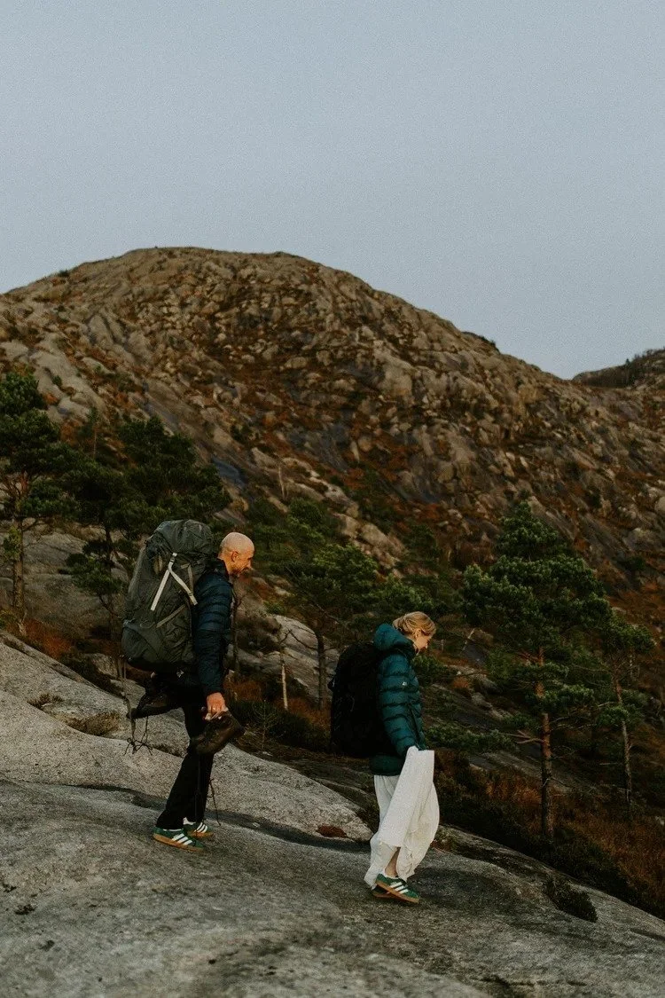 To turgåere på en steinete fjellsti med trær rundt. En mann med sekk og fjellsko, og en kvinne med sekk, jakke og brudekjole