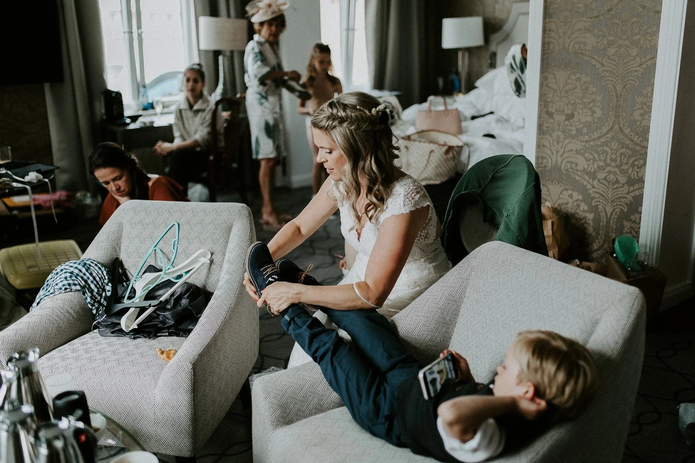 En brud i hvit blondekjole hjelper brudesven til å ta på skoene i et hotellrom. Flere kvinner er til stede i rommet.