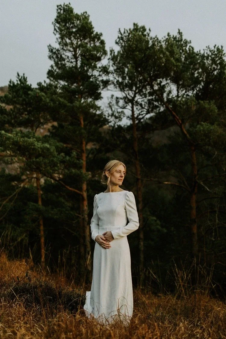 En kvinne i lang hvit kjole står i et felt med høyt gress, omgitt av furutrær i skumringen.