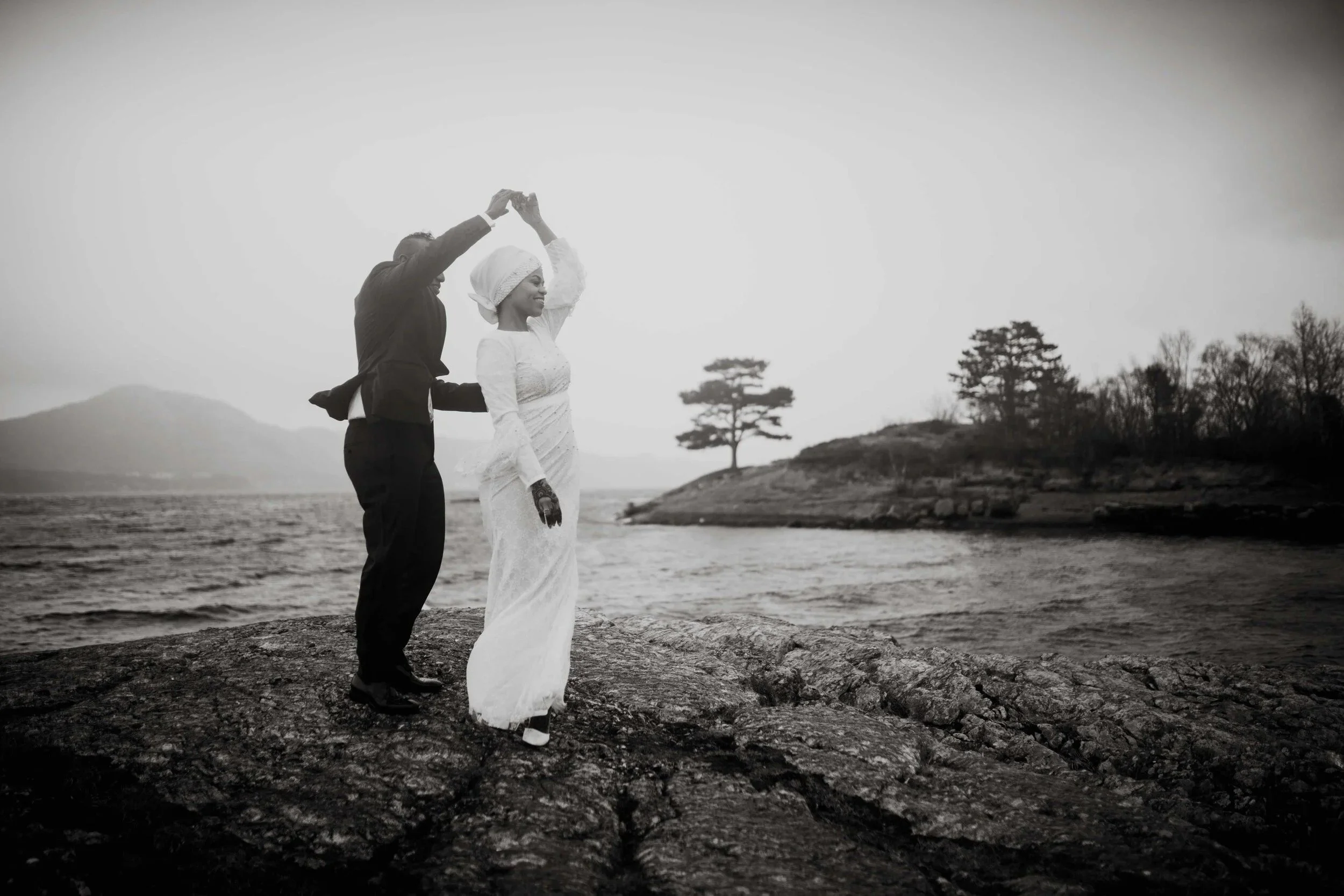 Et par danser på en steinete strand ved vannet, med trær og åser i bakgrunnen, fotografert i sort hvitt.