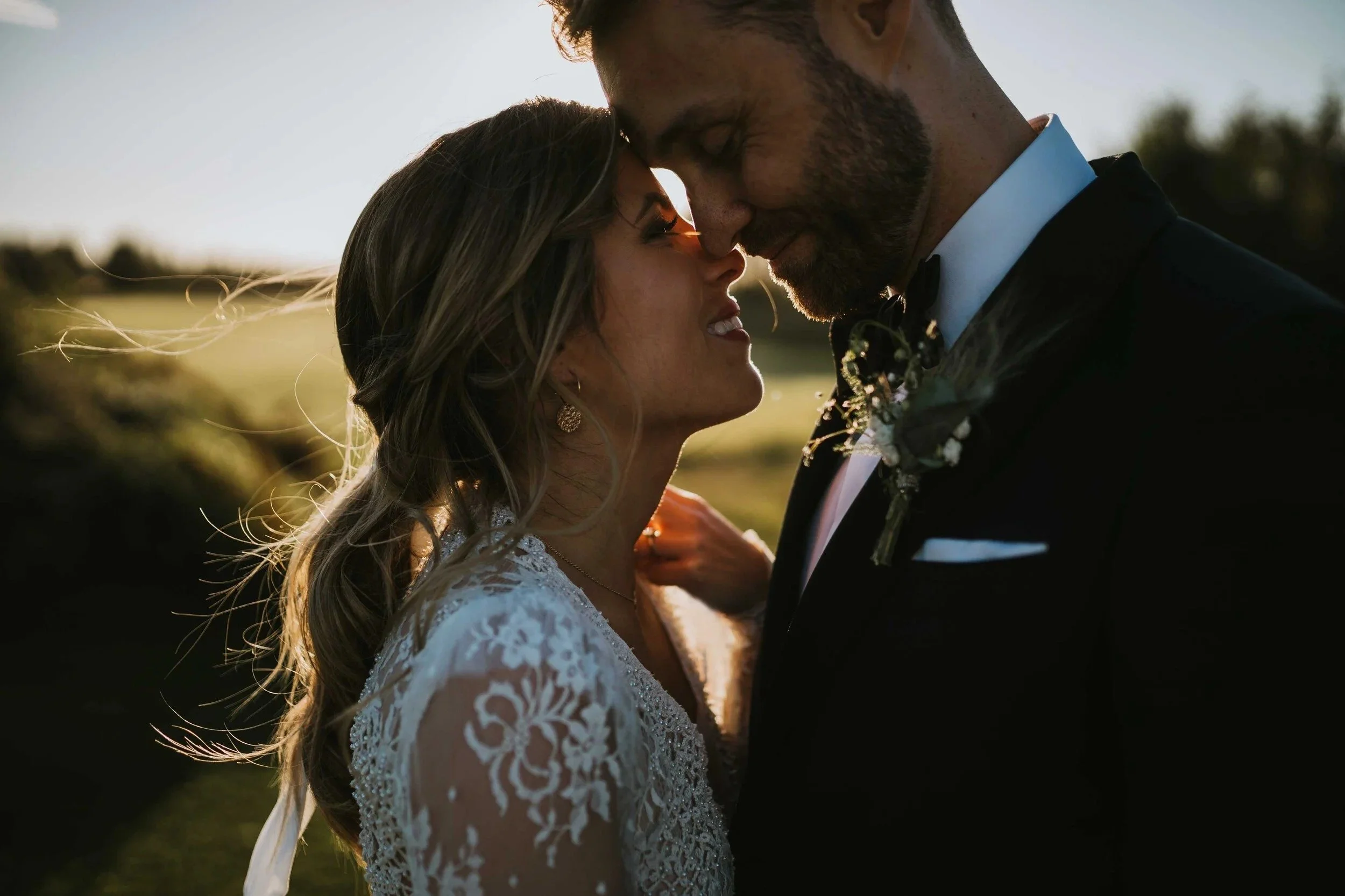 Et brudepar med panner og neser mot hverandre, badet i varmt solnedgangslys under en utendørs bryllupsseremoni.