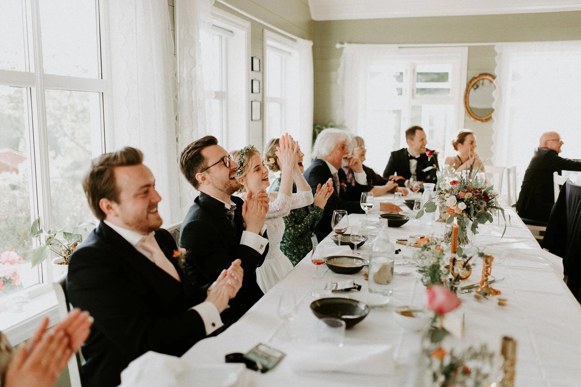 En gruppe mennesker sitter ved et bryllupsbord, smiler og klapper, med blomster og drikke på bordet i et lyst rom med store vinduer.