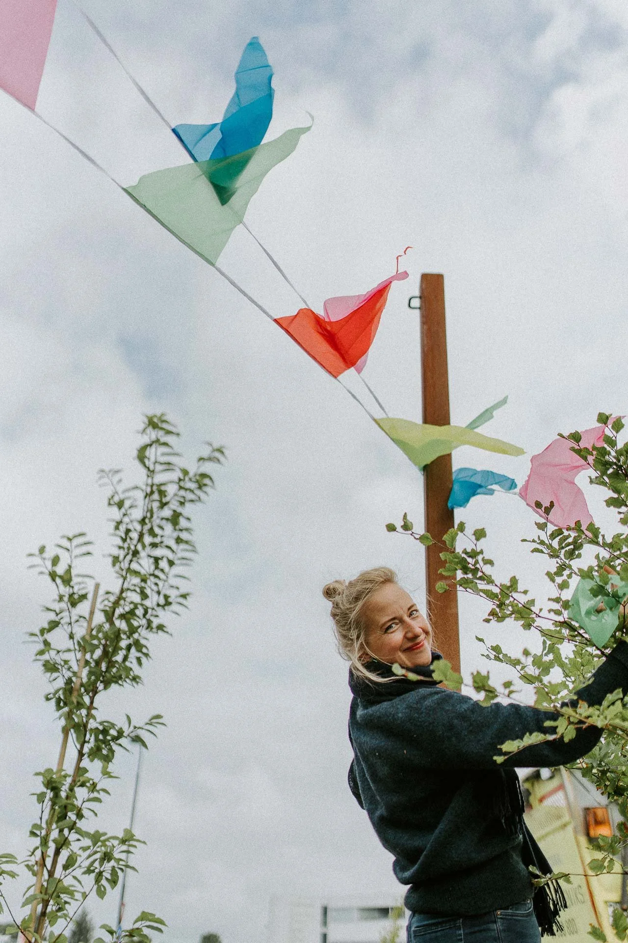 Kvinne som smiler og ser i kamera henger opp vimpler trær og himmel i bakgrunnen naturlig lys, reklamefotografering for stavanger kommune, områdesatsingen i hillevåg