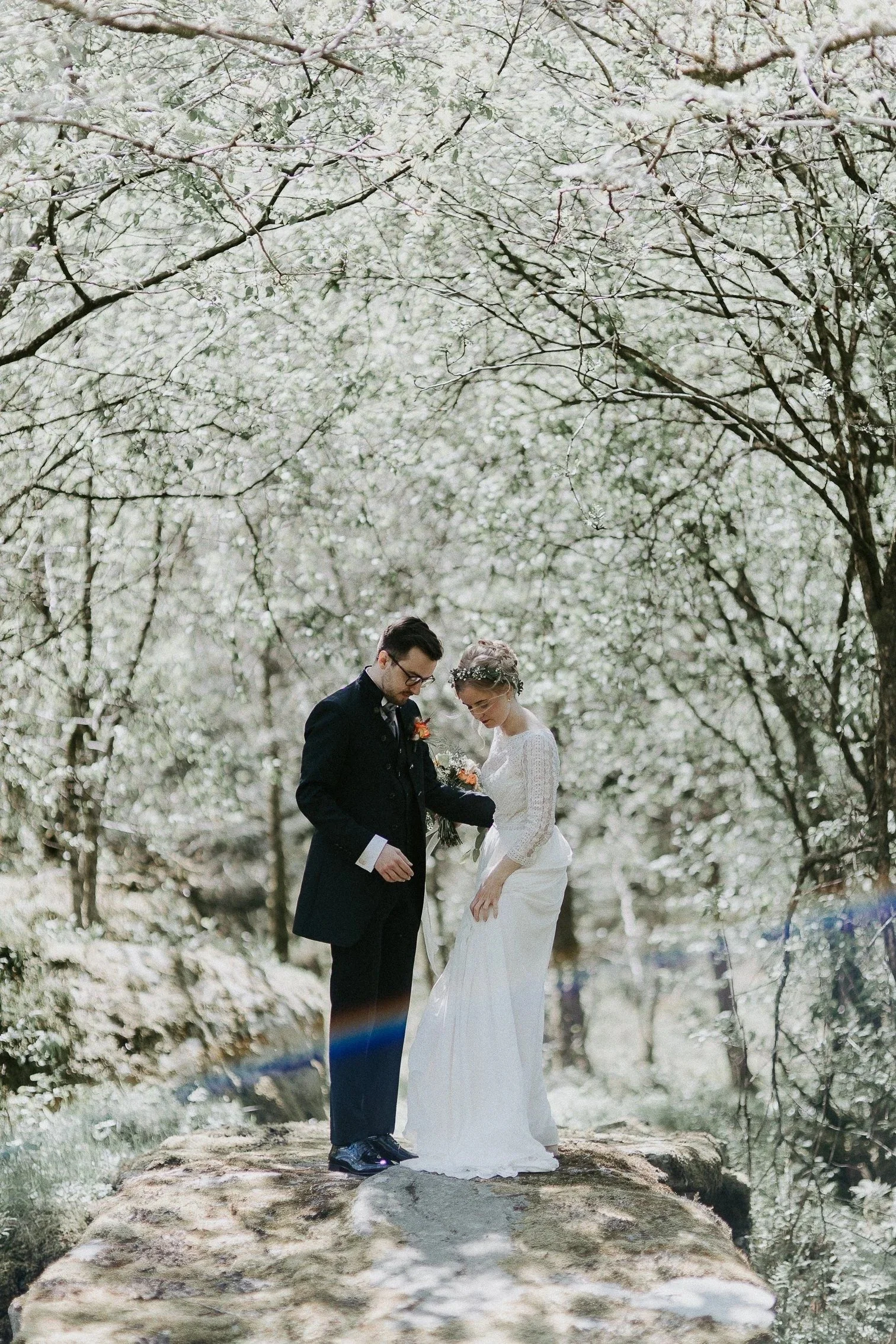 Brud og brudgom står på en stor stein i skogen med blomstrende trær rundt, holder hender og ser ned under fotografering.
