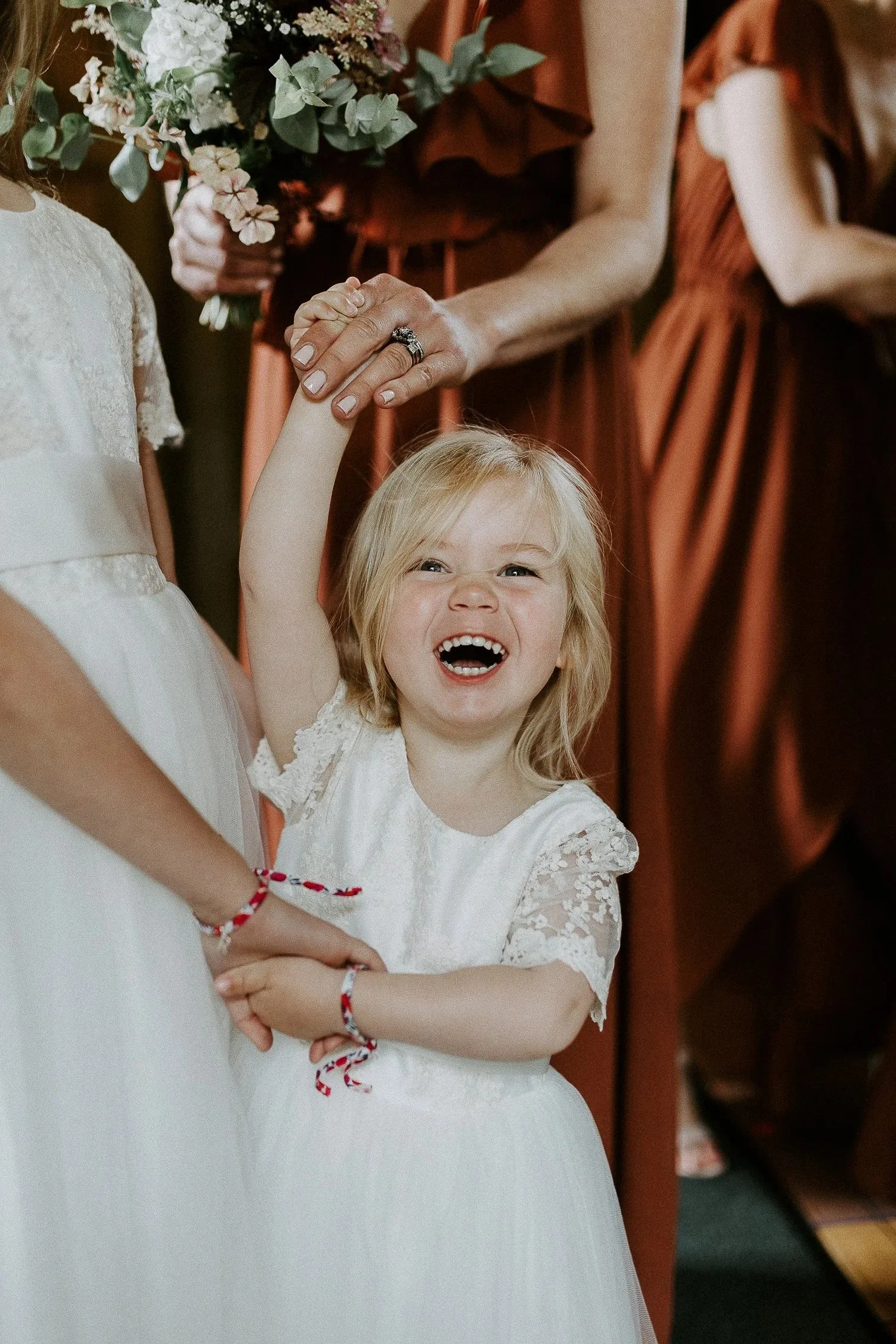 En jente brudepike i hvit blondekjole ler og holder en voksen i hånden. Flere kvinner i rust eller oransje kjoler er synlige i bakgrunnen, én holder en blomsterbukett.
